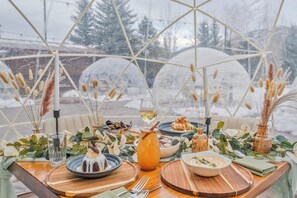 A dining table set with various dishes, a glass of wine, and decorative elements like candles and plants, viewed through a geodesic dome structure.