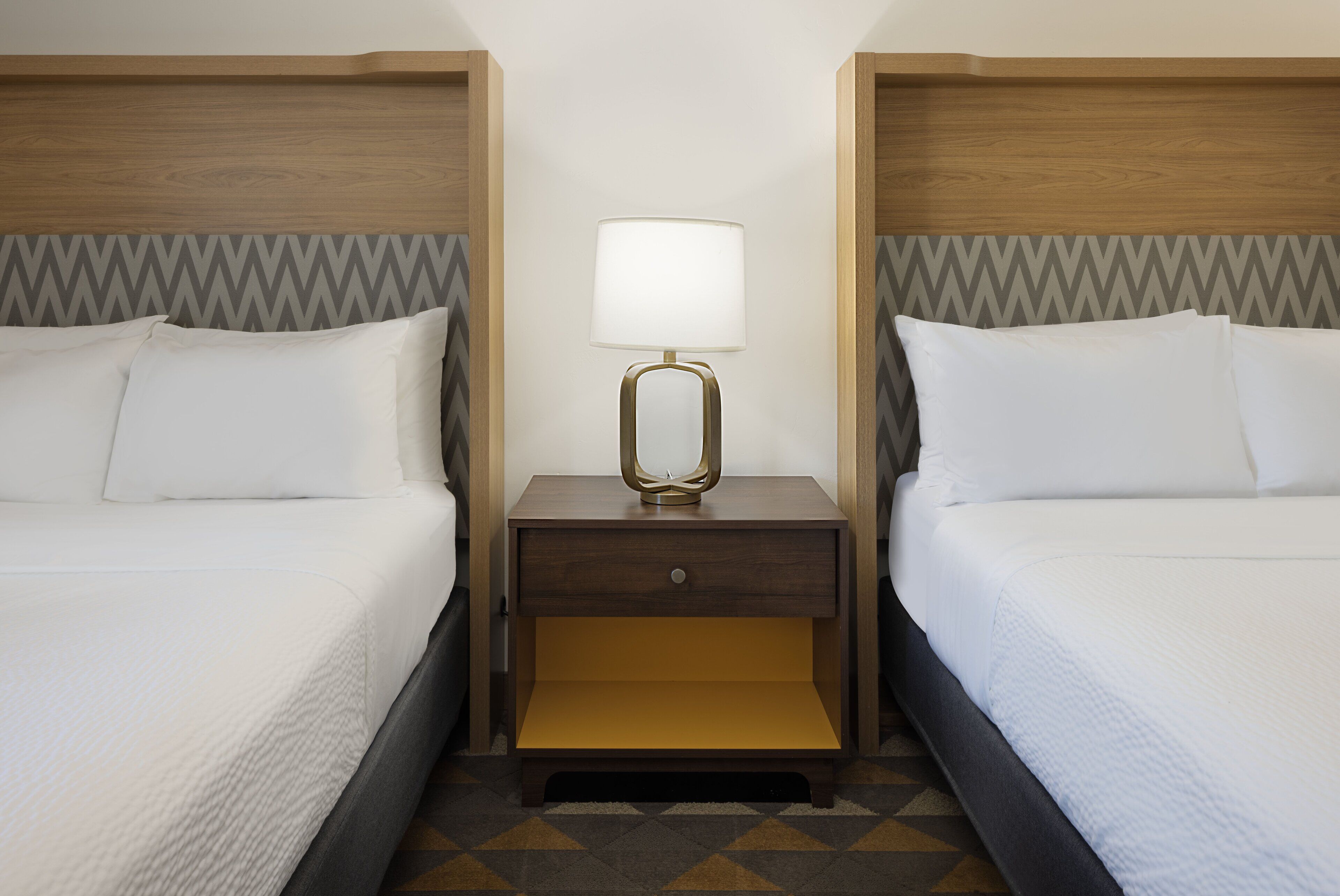 A hotel room with two beds, a nightstand with a lamp, and a patterned floor.