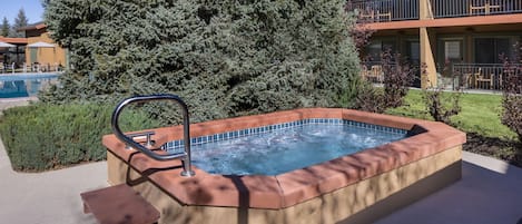 Ein Whirlpool mit Metallleiter, umgeben von einer Betonplattform, einem Schwimmbad und einer großen Nadelbaumart.