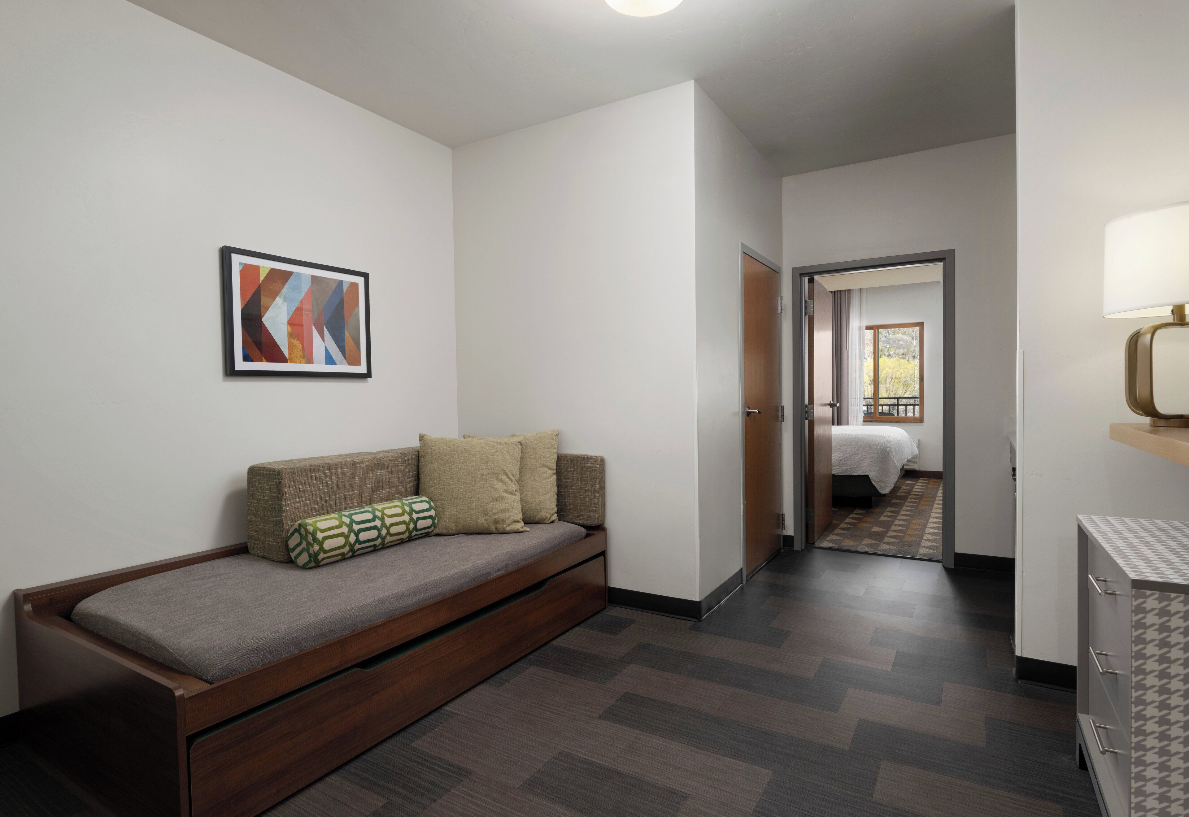 A modern hotel room with a wooden sofa, a framed abstract painting, and a view into the adjacent bedroom.