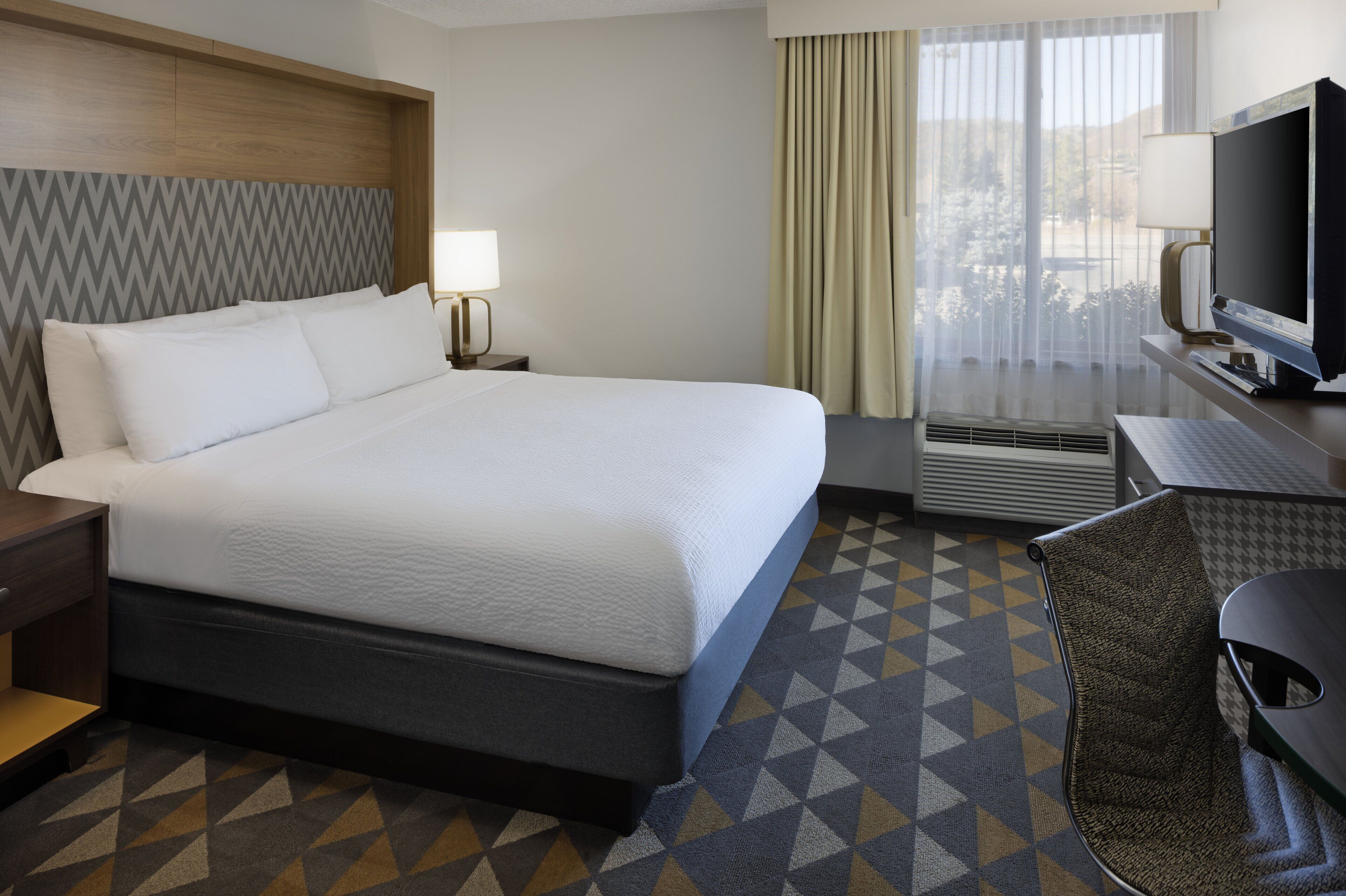 Una habitación de hotel con una cama grande, un escritorio con televisor, una silla y una ventana con cortinas.