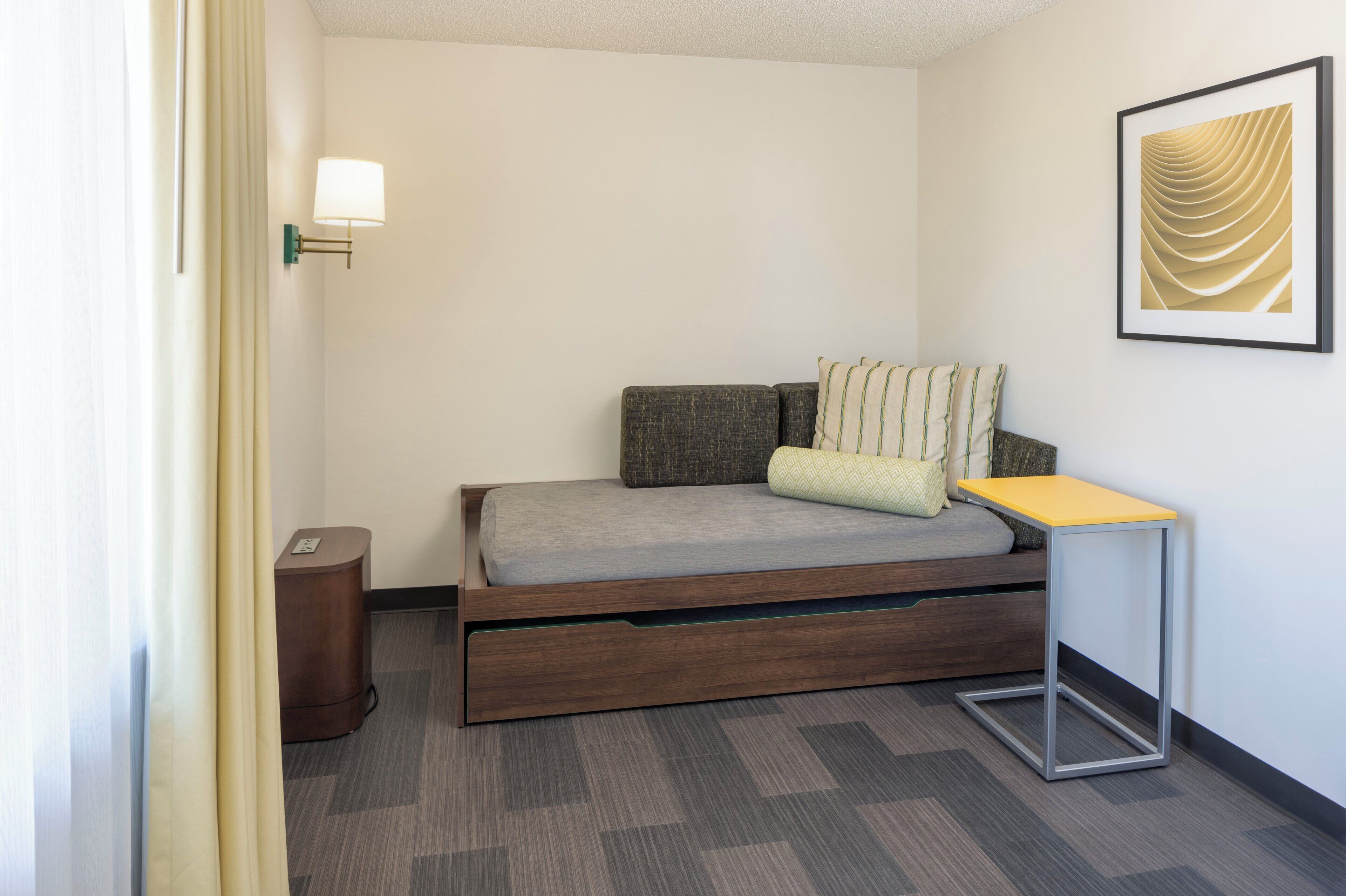 A compact hotel room with a single bed, a small bedside table, a wall-mounted lamp, and a framed artwork.