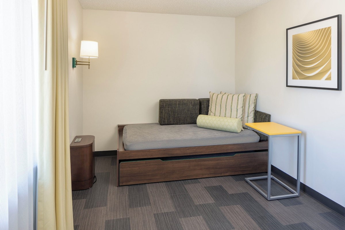 A compact hotel room with a single bed, a small bedside table, a wall-mounted lamp, and a framed artwork.