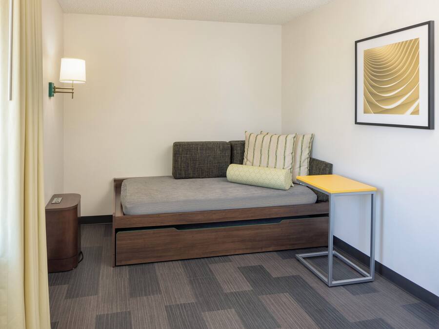A compact hotel room with a single bed, a small bedside table, a wall-mounted lamp, and a framed artwork.