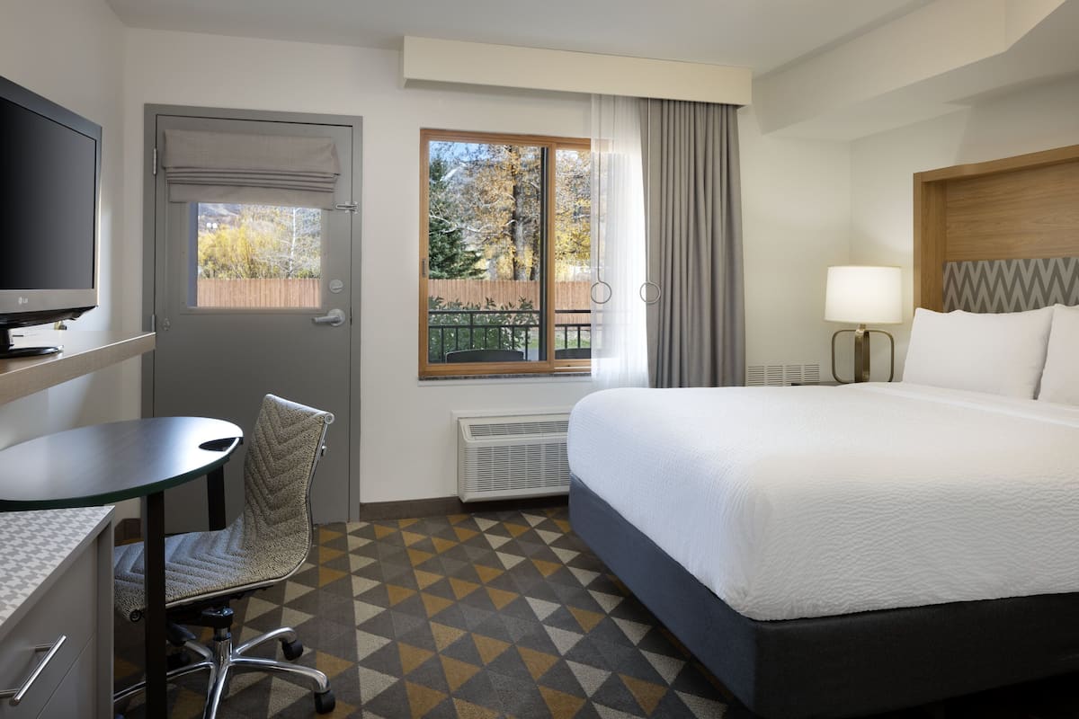 Habitación de hotel con cama, escritorio, silla, televisor, ventana con cortinas y vistas al exterior.