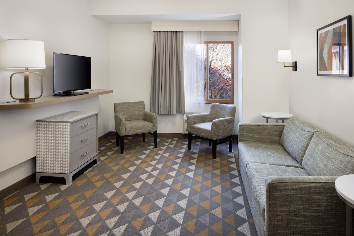 Una habitación de hotel con sofá, dos sillones, televisor sobre un mueble, una mesita, lámpara y piso con diseño.