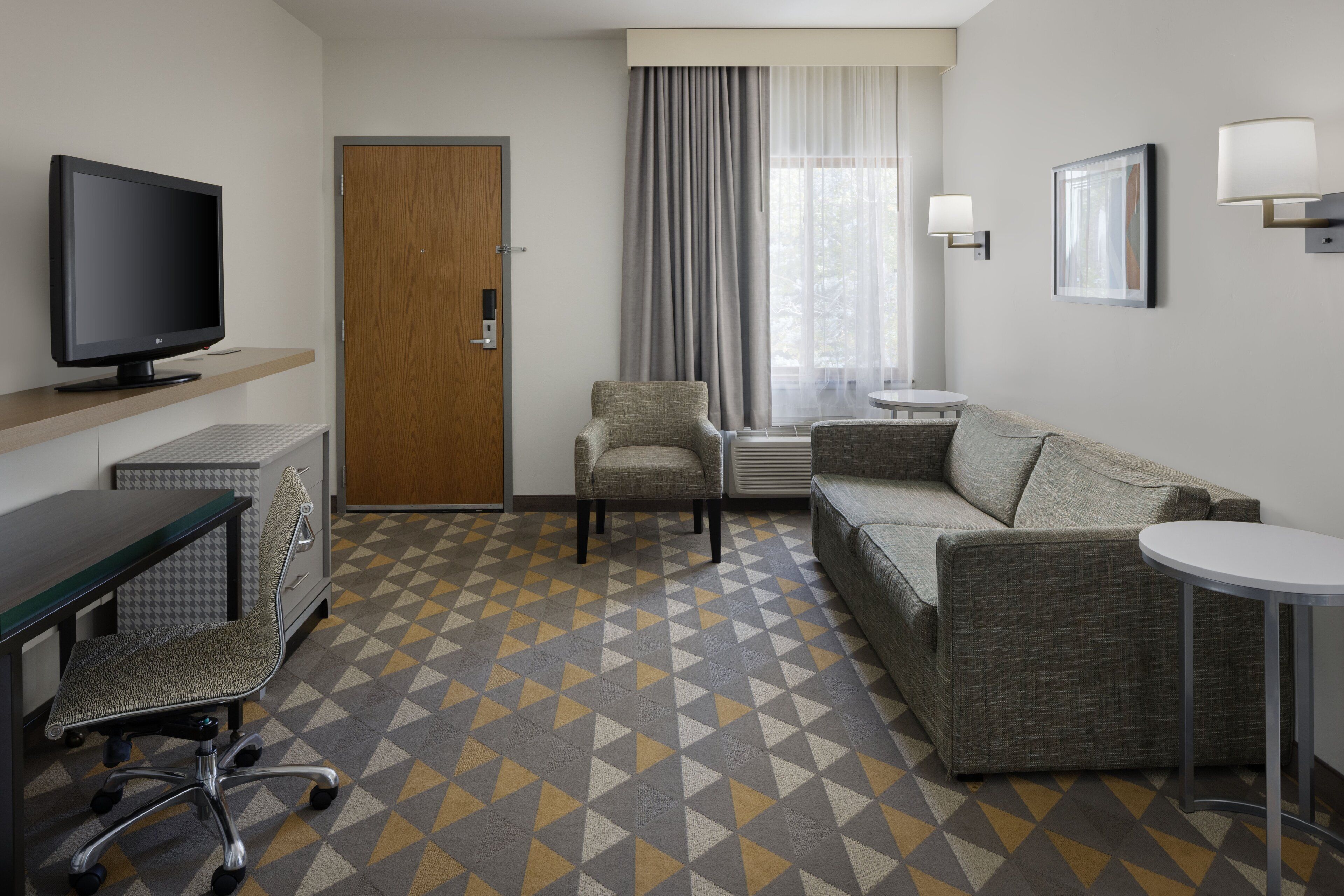 A hotel room with a TV, a sofa, a chair, a small table, and a patterned floor.