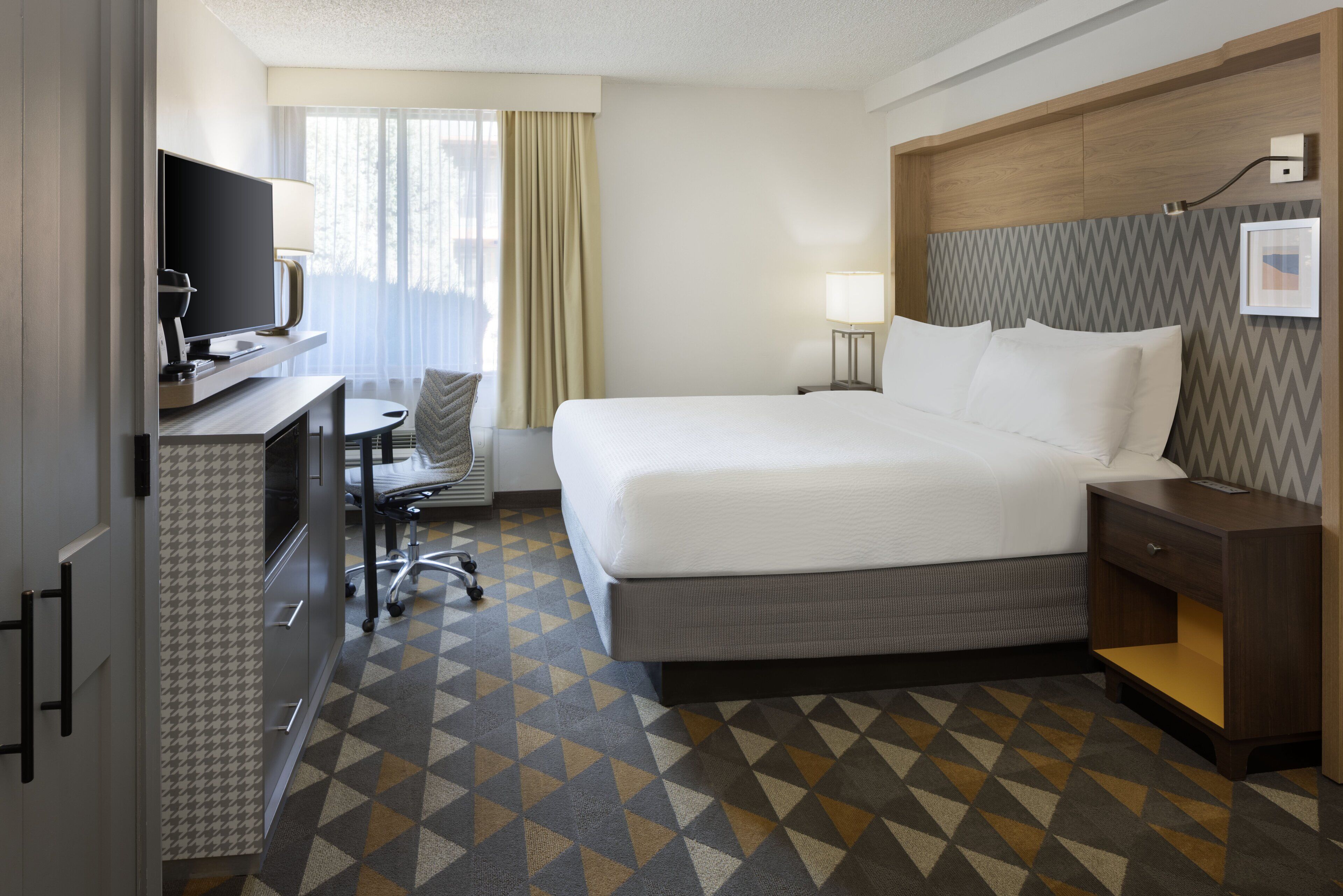 A hotel room with a bed, a desk with a TV, a chair, a lamp, and a patterned floor.