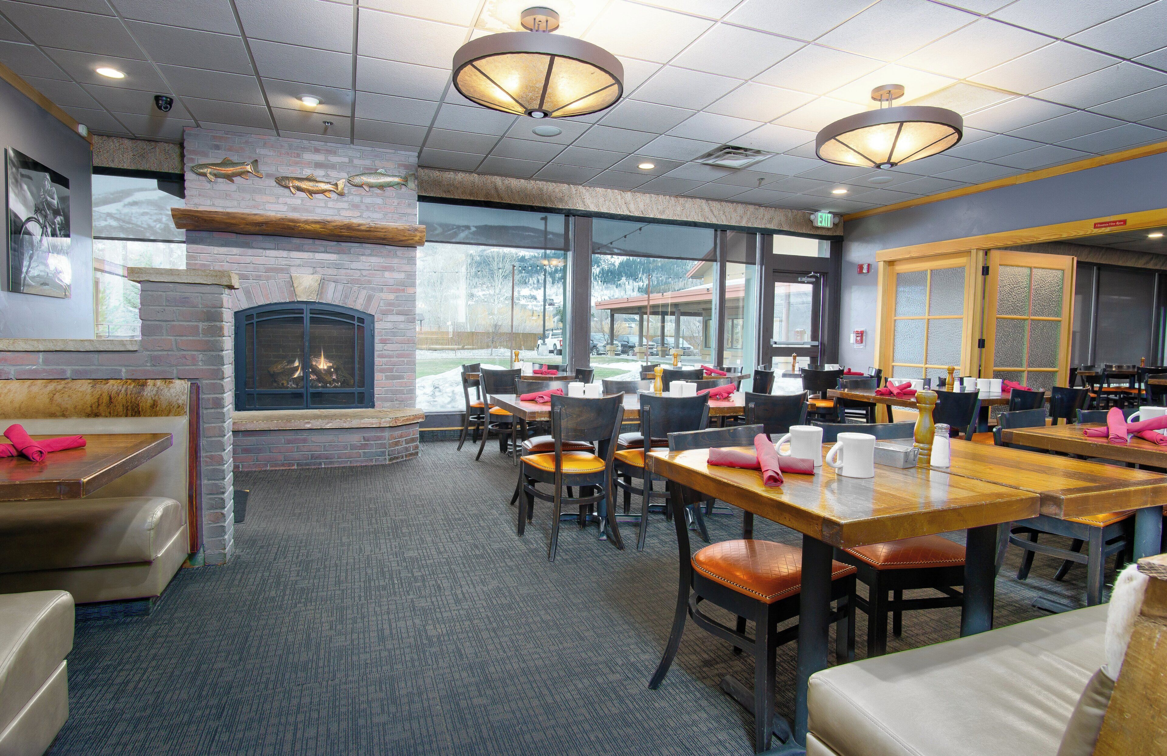 A restaurant with brick walls, a fireplace, and large windows. Tables and chairs are arranged for dining.