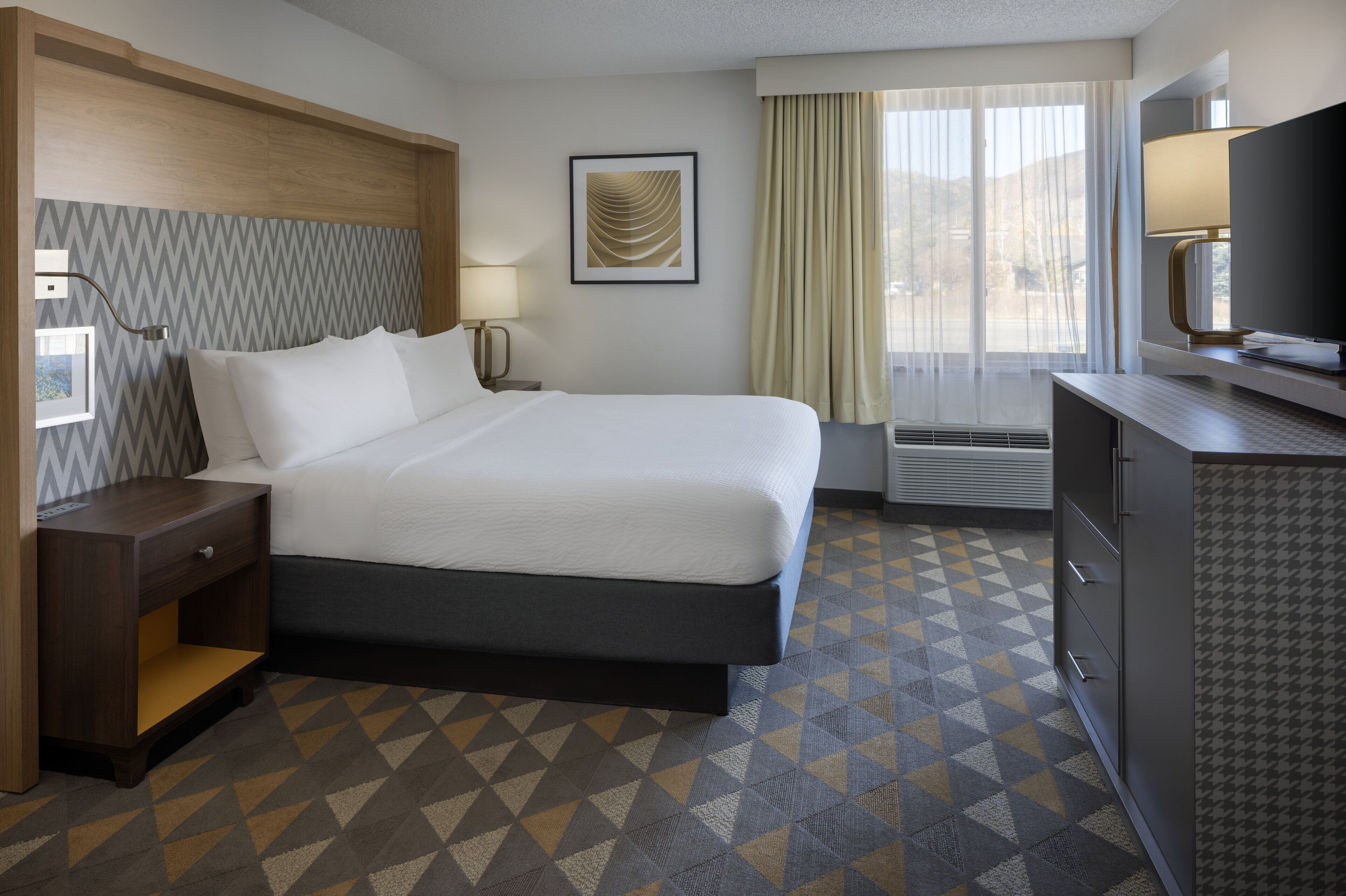 Una habitación de hotel con cama, mesita de noche, escritorio, televisión y ventana con cortinas.