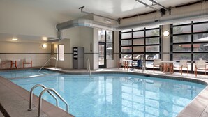 An indoor swimming pool with clear water, surrounded by lounge chairs and tables, large windows, and visible air ducts.