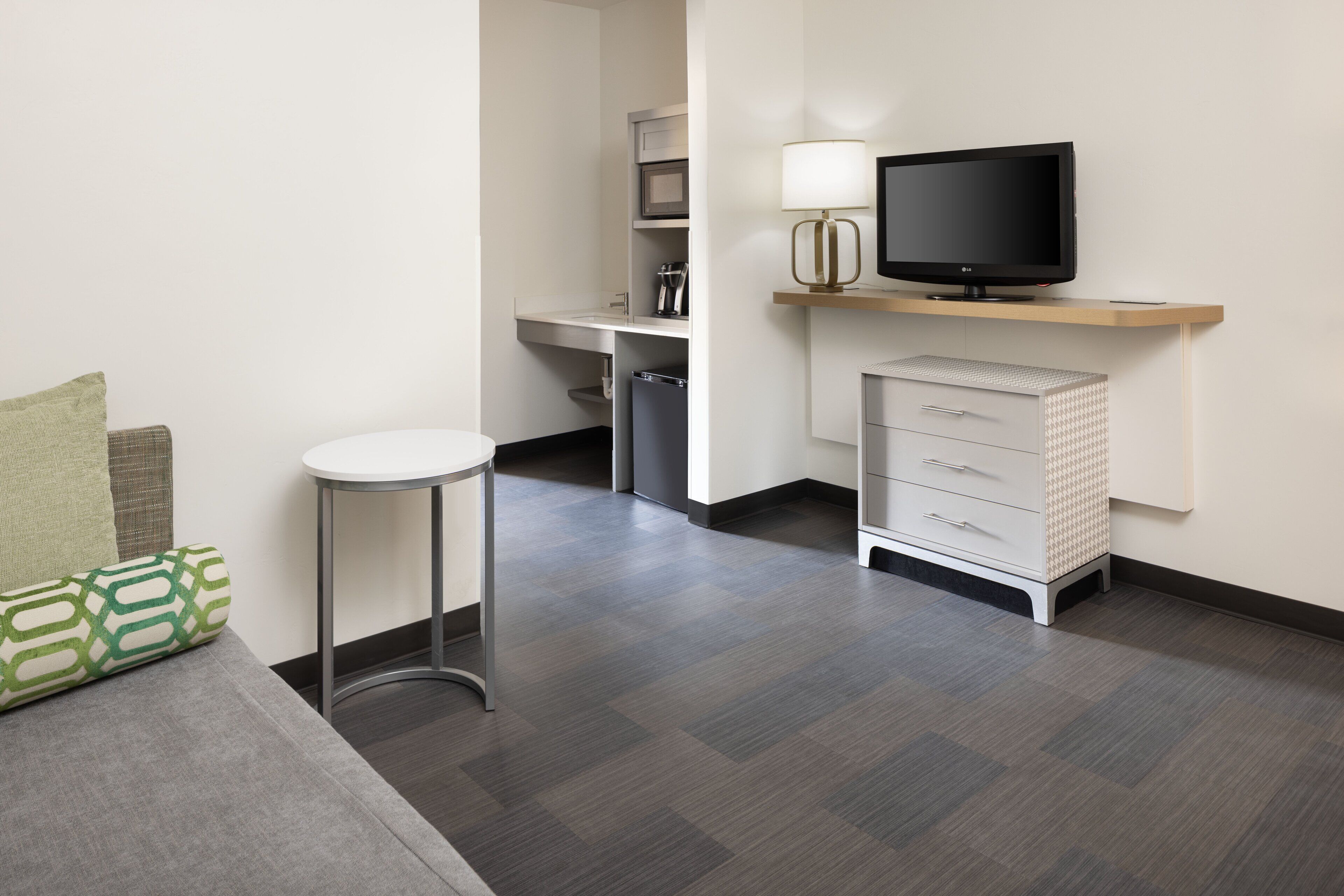 A modern hotel room with a grey sofa, a small round table, a TV on a wooden shelf, a desk with a chair, and a cabinet with drawers.