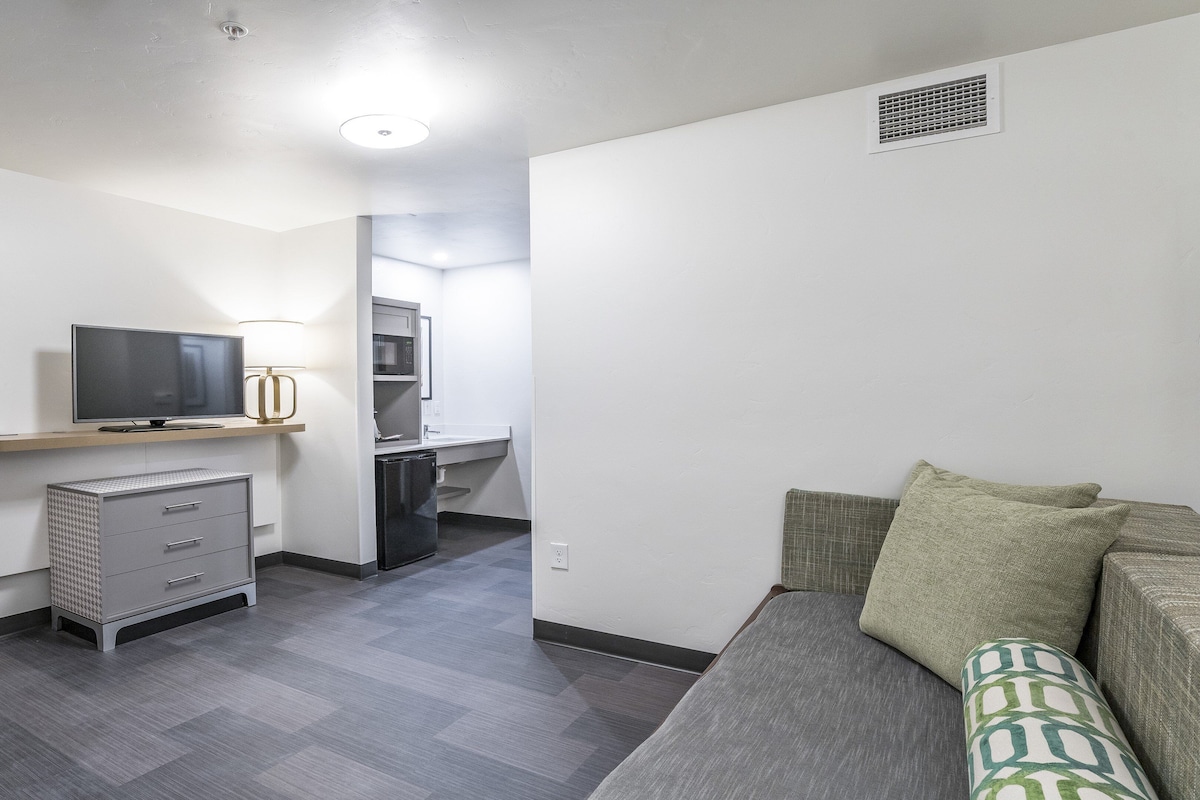 A compact living space with a kitchenette, a flat-screen TV, a small refrigerator, and a sofa with patterned cushions.