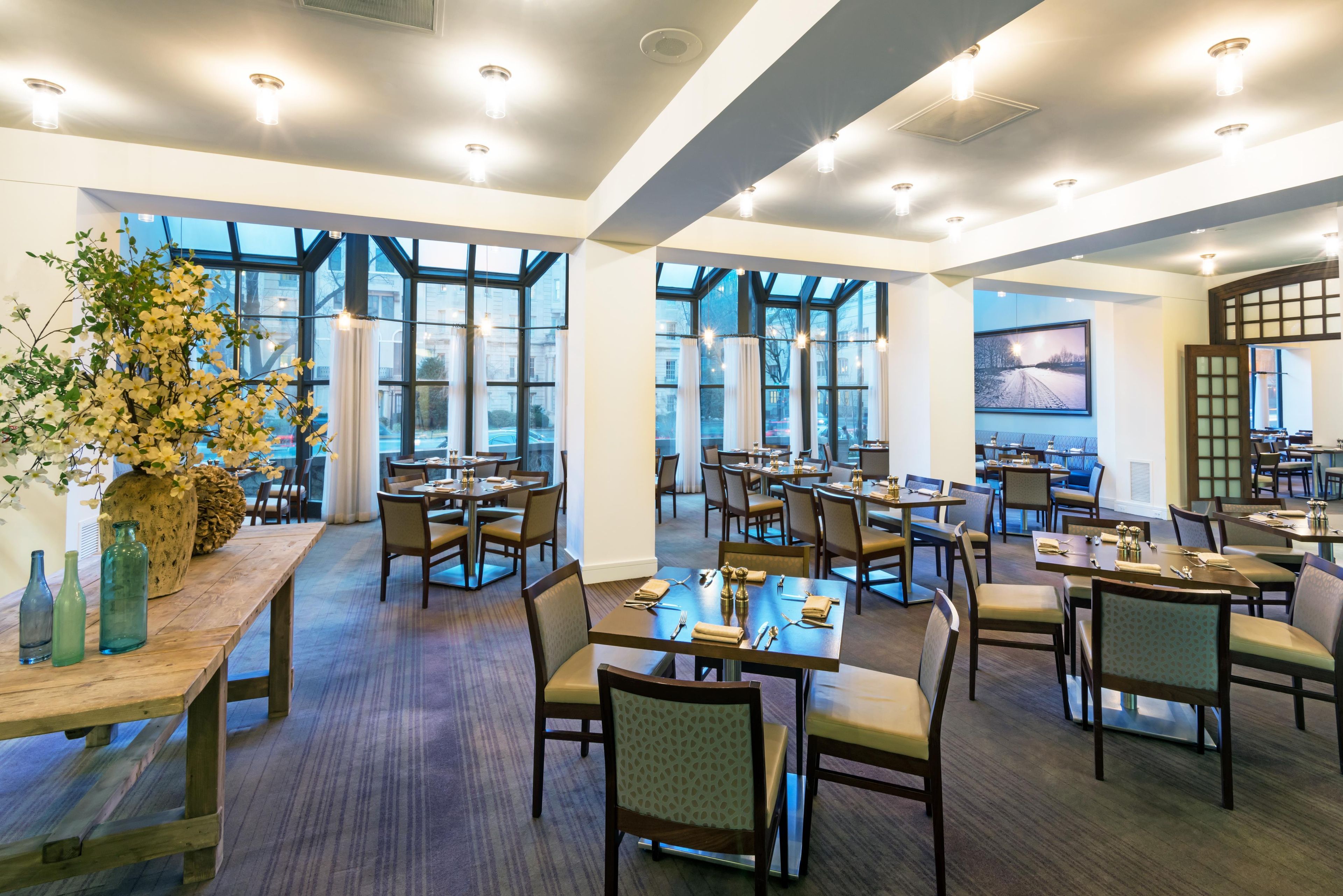 A spacious dining area with multiple tables and chairs, large windows offering views, and a decorative plant arrangement.
