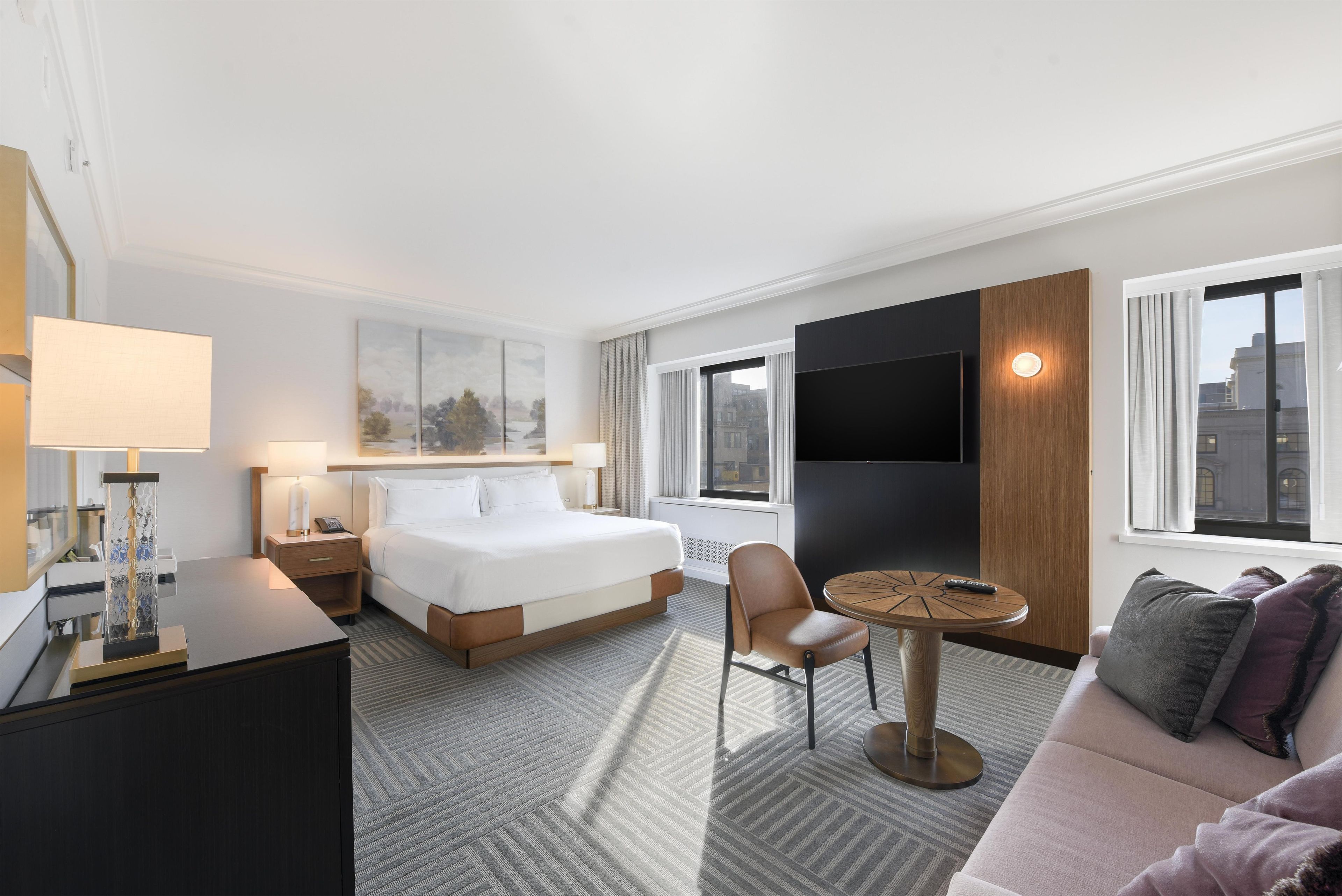 Ett hotellrum med en säng, ett skrivbord, en stol, en soffa, en tv och ett fönster med utsikt över byggnader.
