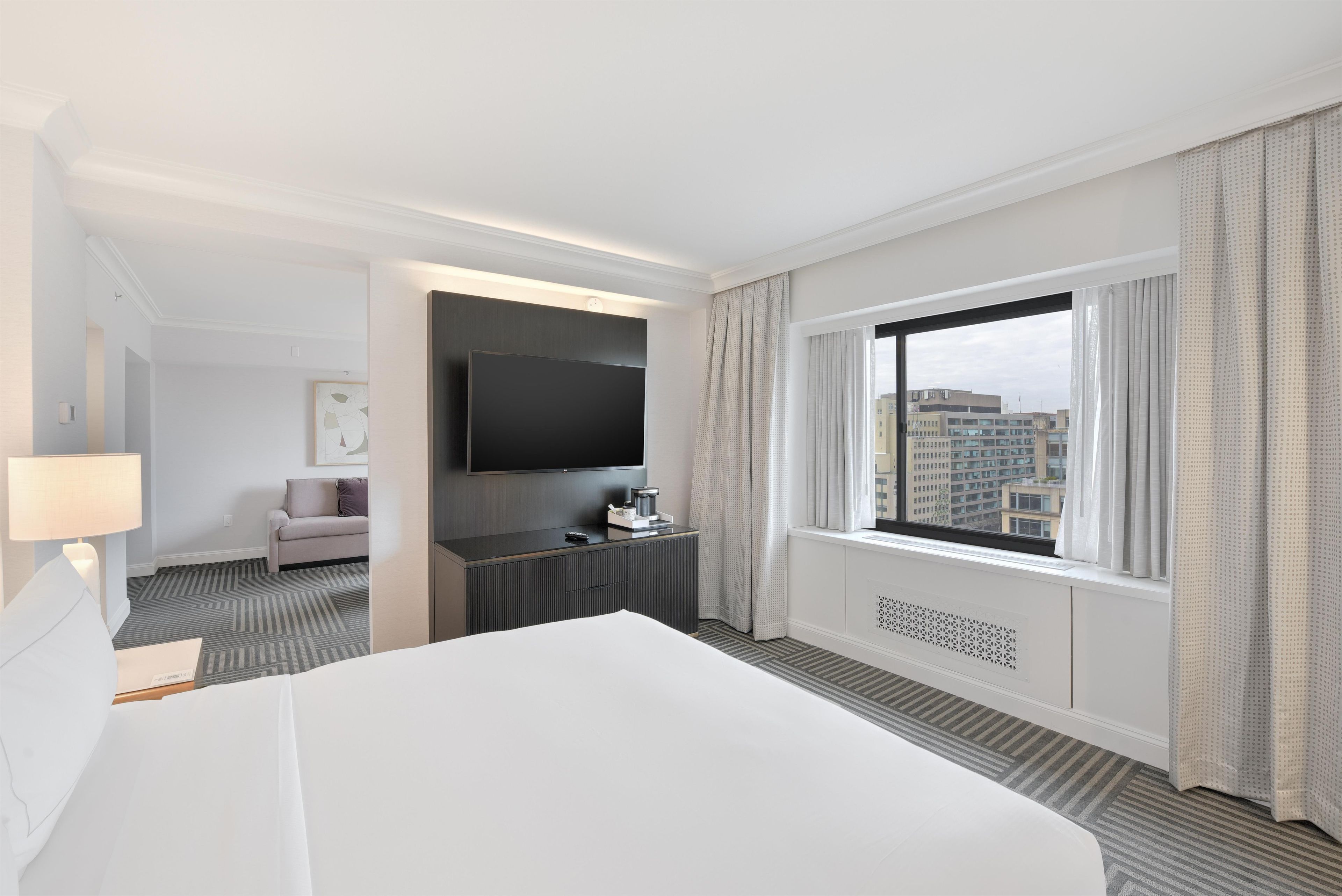 A hotel room with a large bed, a flat-screen TV, a desk with a coffee maker, a sofa, and a window with curtains.