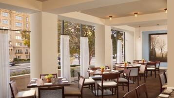 A restaurant with multiple tables and chairs, large windows with curtains, and a view of a street and buildings.