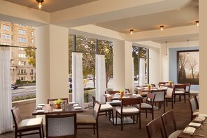 A restaurant with multiple tables and chairs, large windows with curtains, and a view of a street and buildings. - Capital Hilton (Washington)