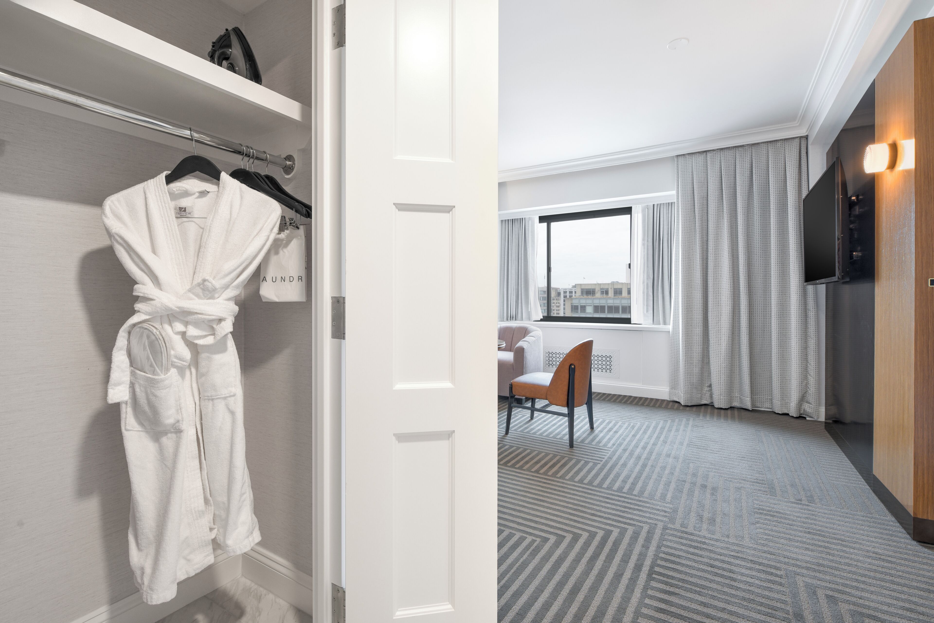 A hotel room with a robe hanging on a rack, a dining area with a chair, and a large window with curtains.