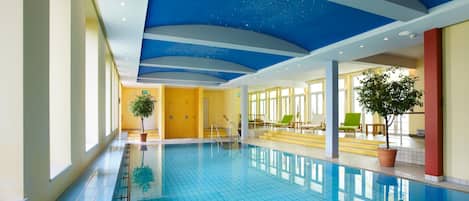 An indoor swimming pool with clear blue tiles, a starry ceiling design, and a sunlit room with seating and plants.