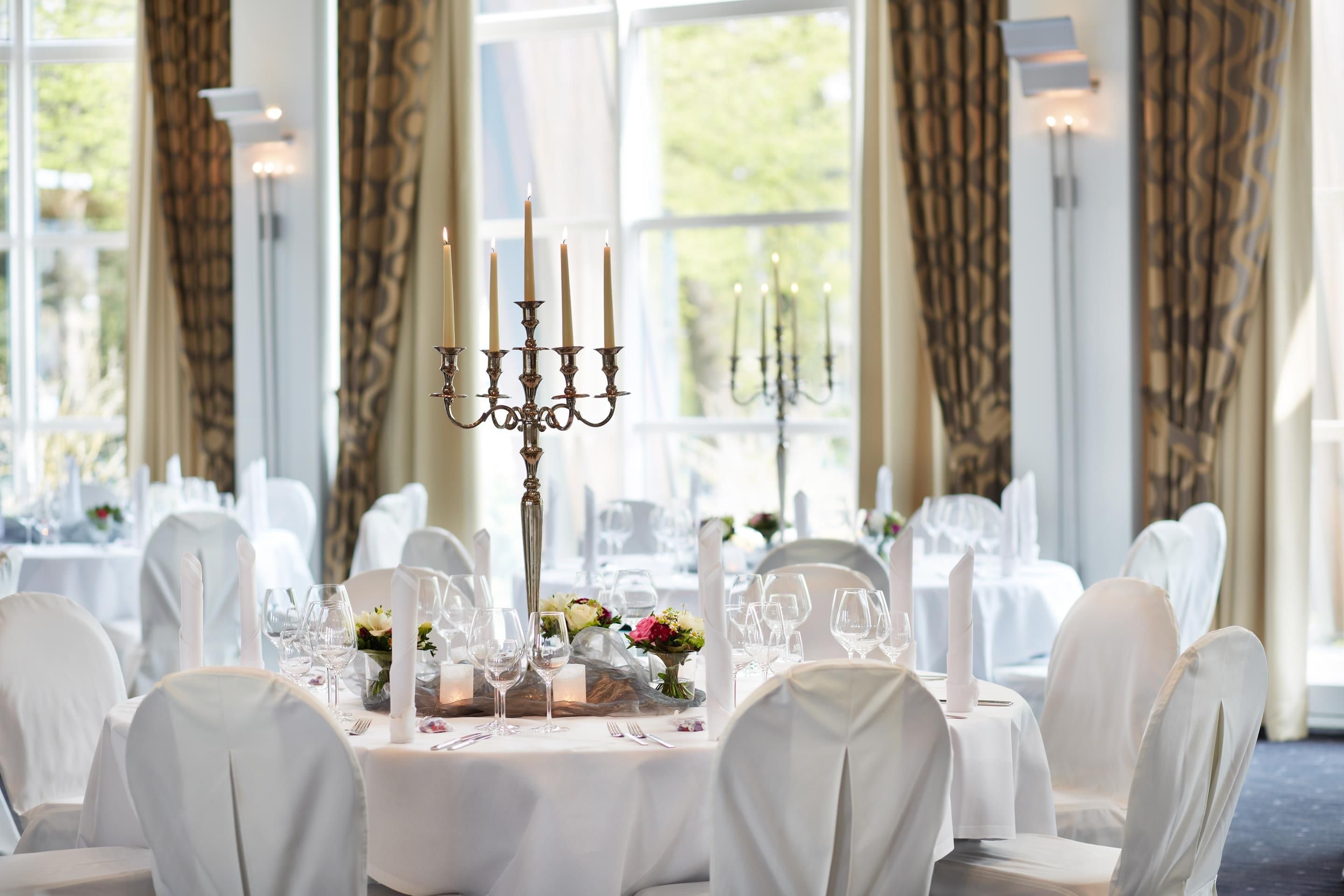 A formal dining room with tables set for a meal, featuring candelabras and floral centerpieces.