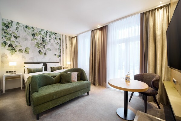 Habitación de hotel con cama, sofá verde, una mesa redonda, una silla, televisor y una pared con estampado de hojas.
