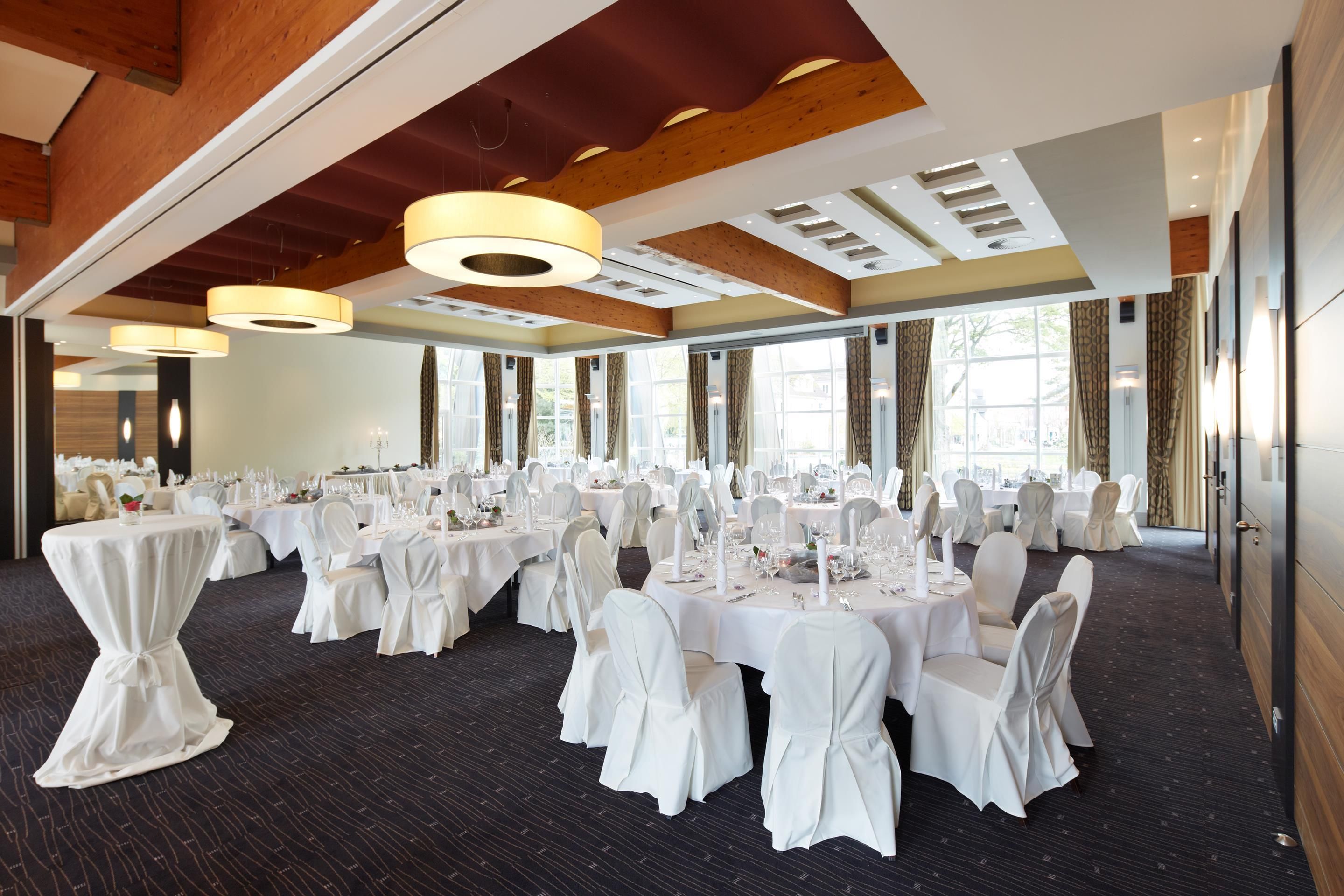 Un salón de banquetes con mesas redondas dispuestas para comer, ventanas amplias y un techo alto con vigas de madera.