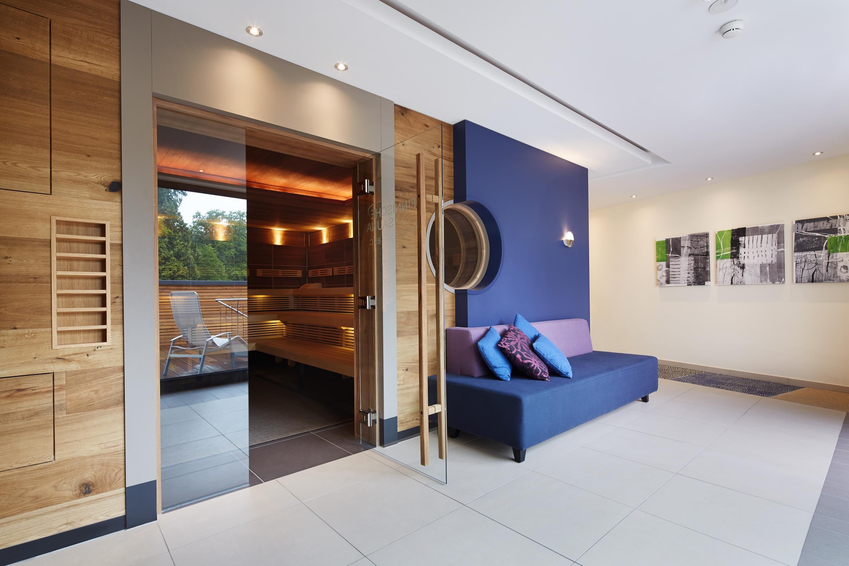Ein modernes Interieur mit Glastür zum Saunabereich, einem blauen Sofa mit Kissen und einem runden, an der Wand befestigten Spiegel.