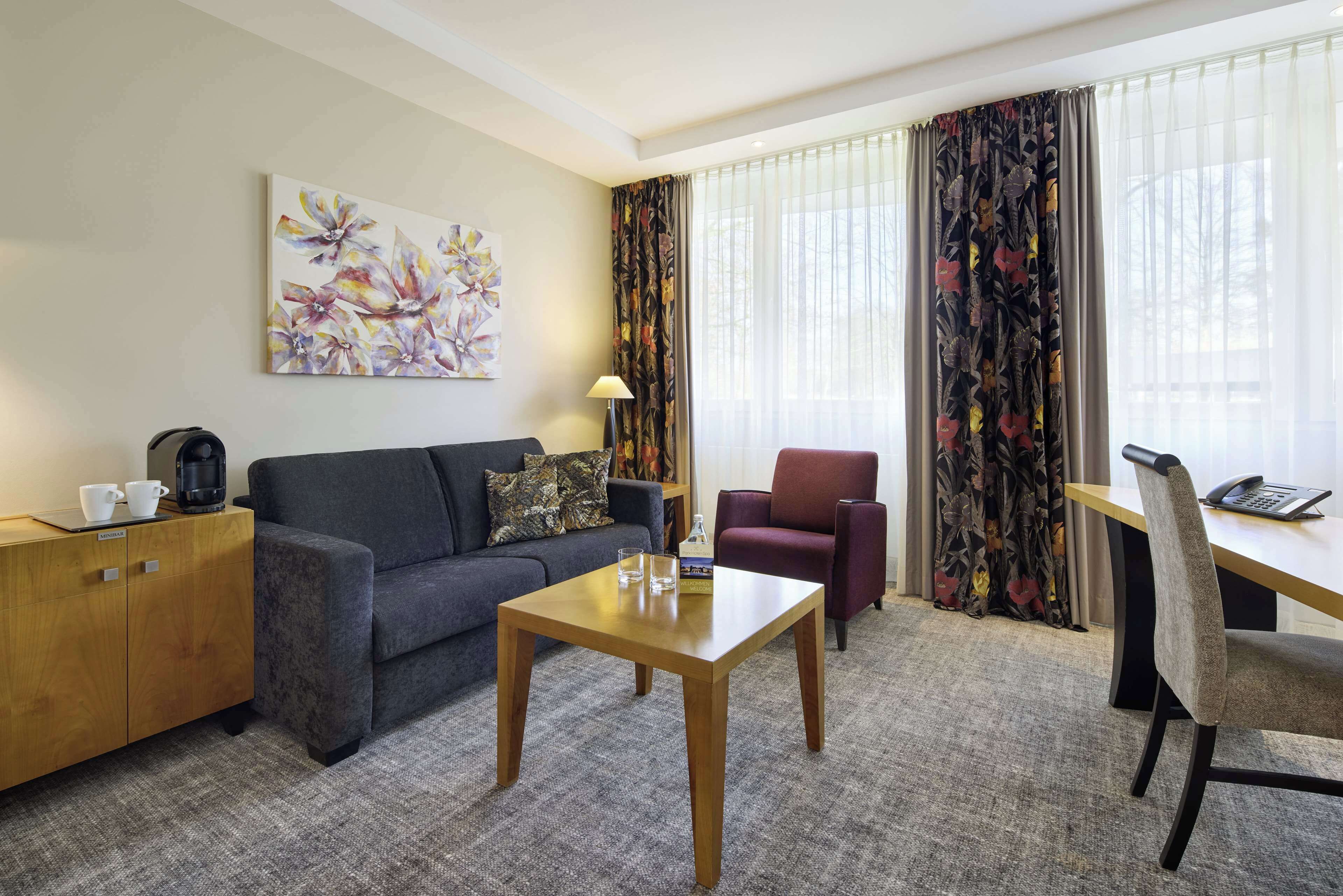 Habitación de hotel con sofá, una mesita de madera, un escritorio con silla, una lámpara y un cuadro en la pared.