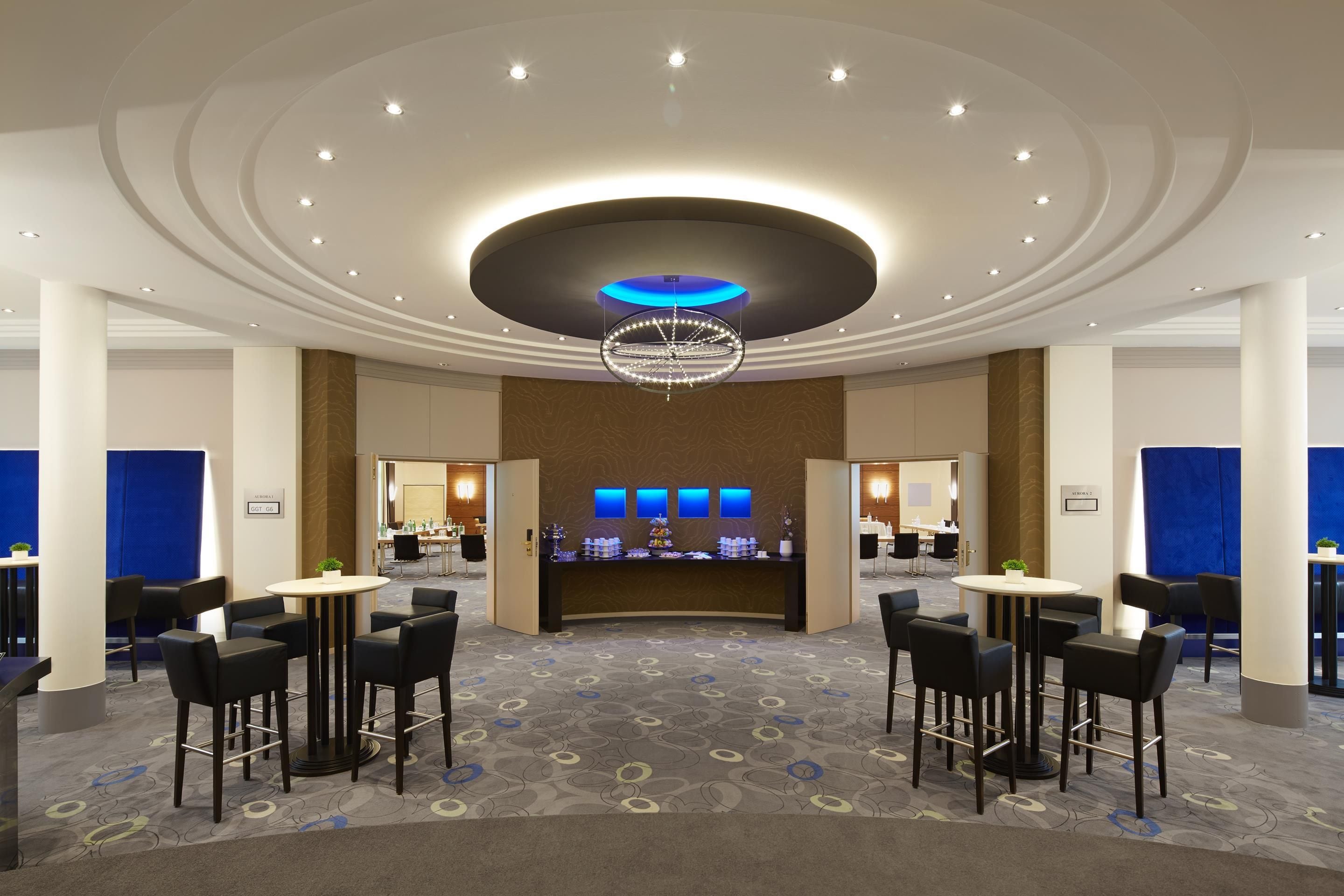 Un vestíbulo de hotel con una zona de bar central, diversas opciones de asientos y un diseño de techo distintivo.