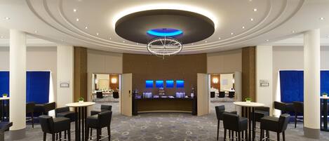 Un vestíbulo de hotel con una zona de bar central, diversas opciones de asientos y un diseño de techo distintivo.