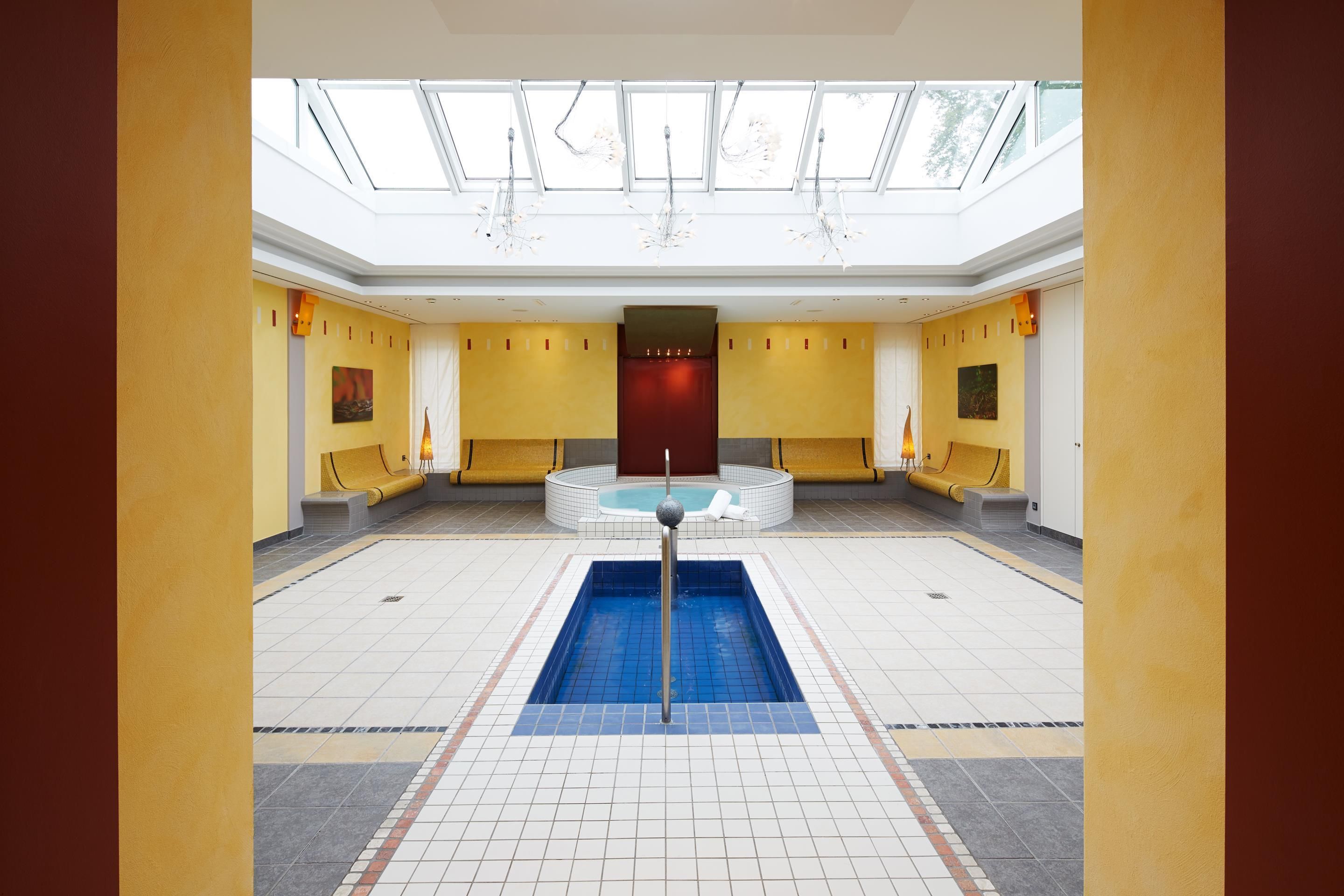 Une grande piscine intérieure dotée d’un jacuzzi central, entourée d’espaces de détente et surmontée d’un haut plafond agrémenté de lucarnes.