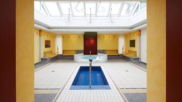 Une grande piscine intérieure dotée d’un jacuzzi central, entourée d’espaces de détente et surmontée d’un haut plafond agrémenté de lucarnes.
