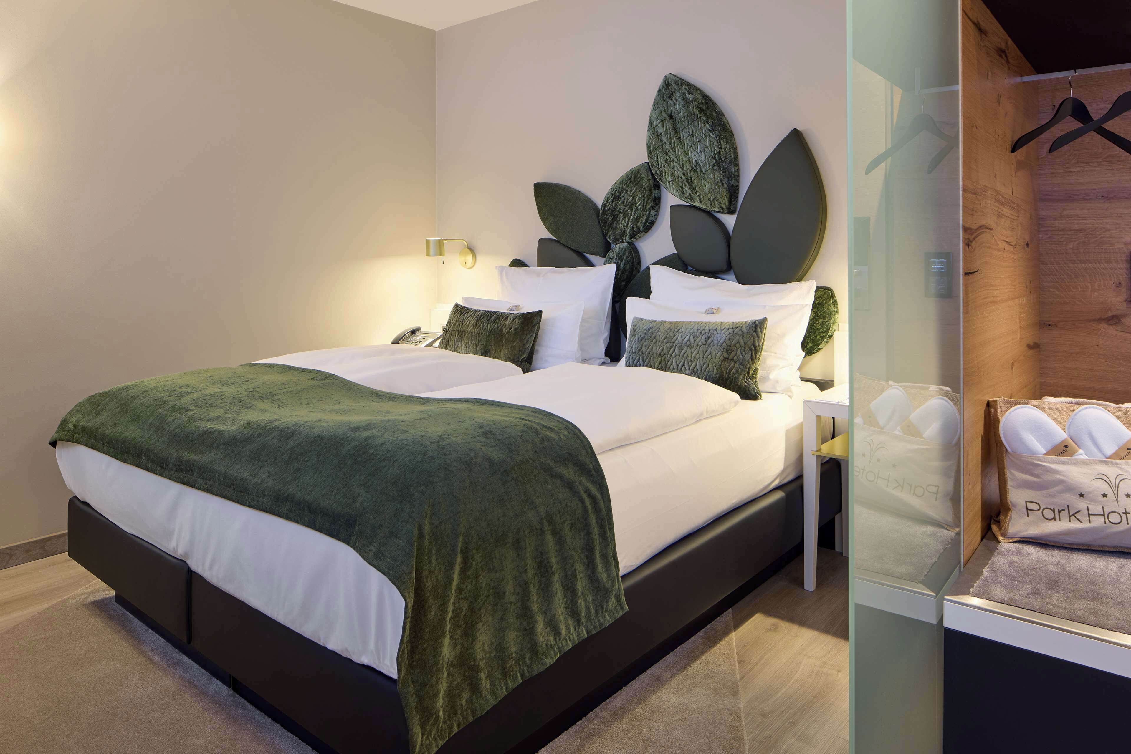A modern hotel room with a large bed, a nightstand, a wall-mounted light, and a wooden panel with a hanging rack.