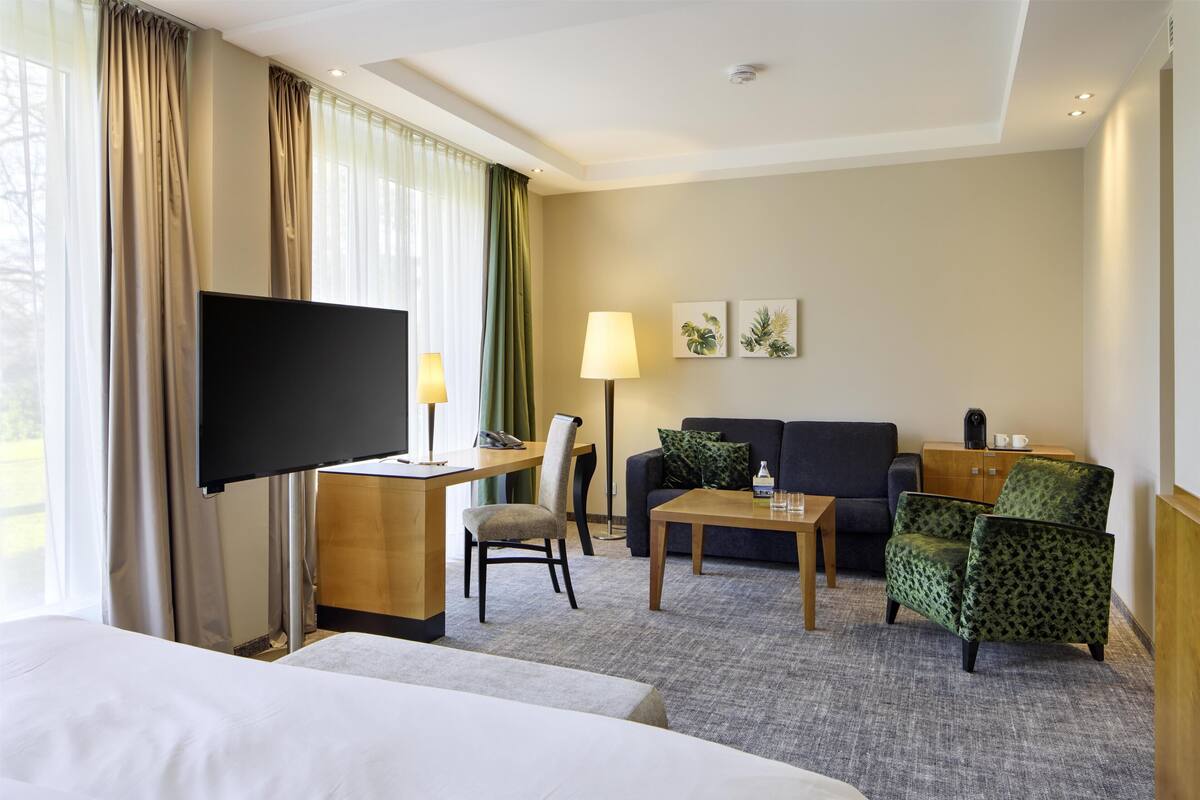 A hotel room with a bed, a desk with a flat-screen TV, a sofa, a chair, a coffee table, and a lamp.