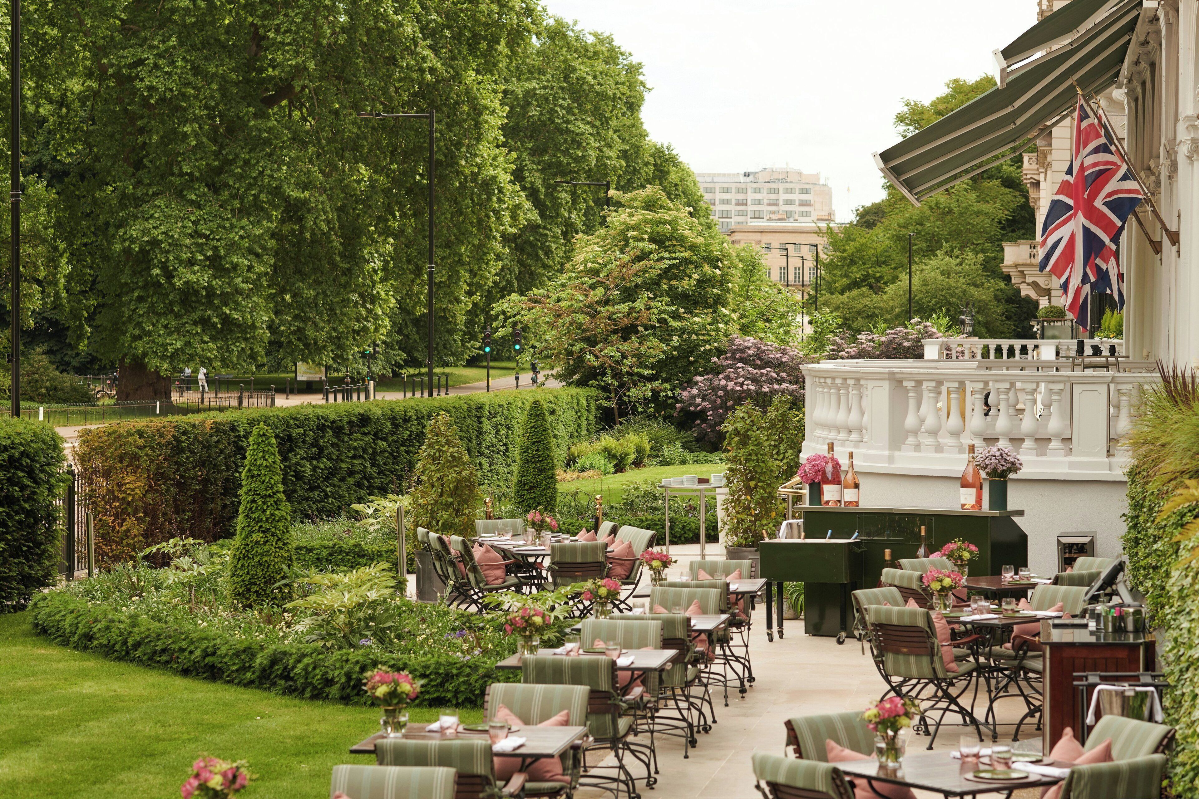 An outdoor dining area with tables and chairs, surrounded by well-maintained hedges and trees.
