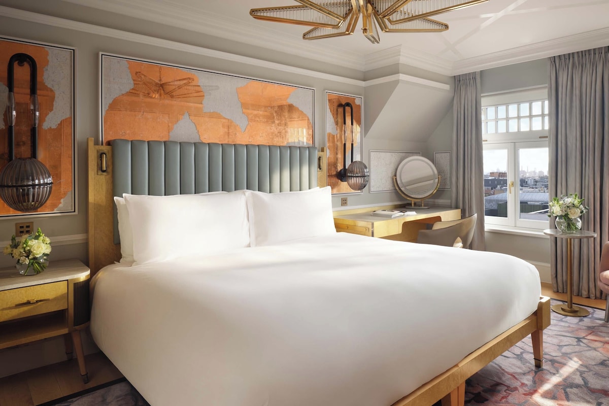 A hotel room with a large bed, a desk, a chair, a large window with a city view, and artwork on the walls.