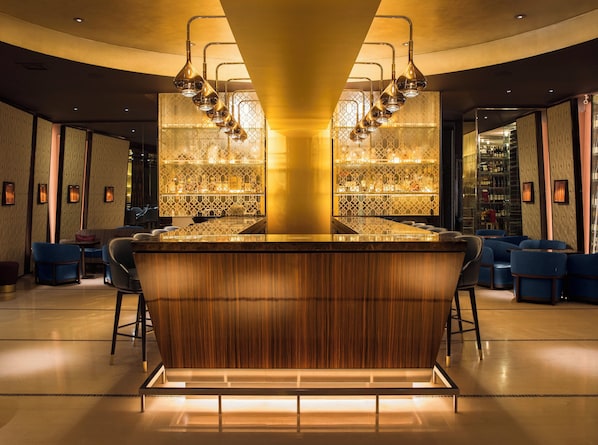A bar area with a central wooden bar, blue seating, and a patterned wall with a bar in the background.