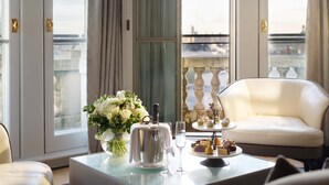 Een luxueuze kamer met balkon, een witte bank, een tafel met bloemdecoratie, een zilveren theepot en een plateau met desserts.