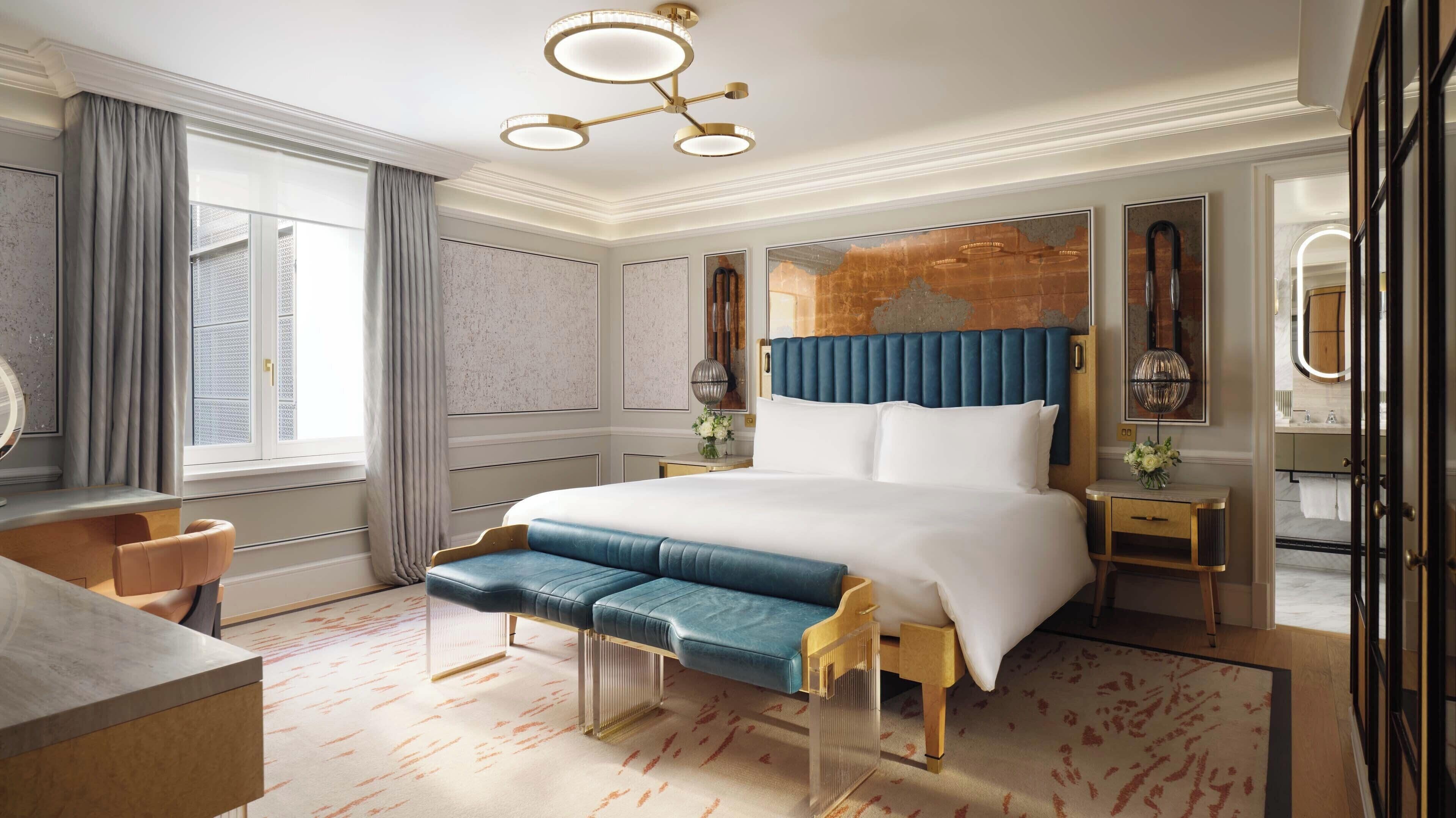 A hotel room with a large bed, a desk, a chair, a vanity with a mirror, and a window with curtains.