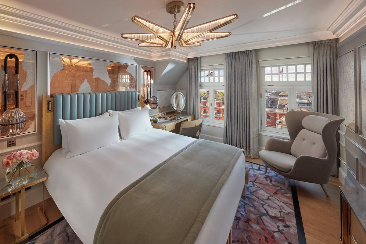 Ein Hotelzimmer mit einem großen Bett, einem Schreibtisch, einem Stuhl, einem Spiegel und Blick auf Gebäude vor dem Fenster.