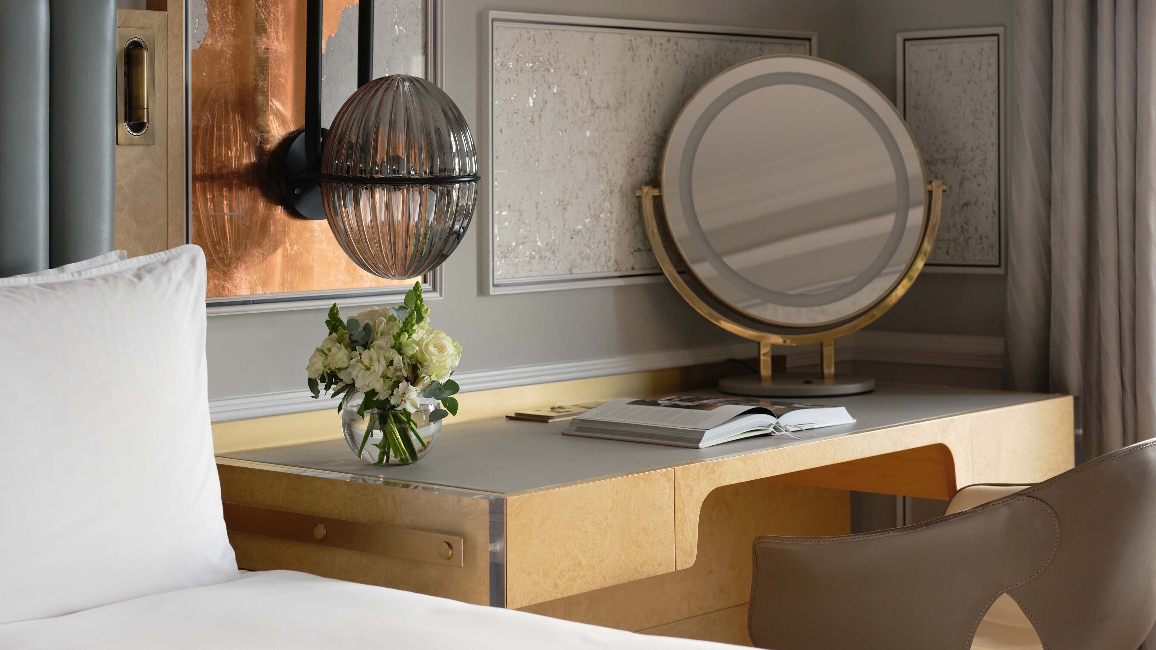 A hotel room with a wooden desk, a round mirror, a vase with flowers, and a bed with white pillows.