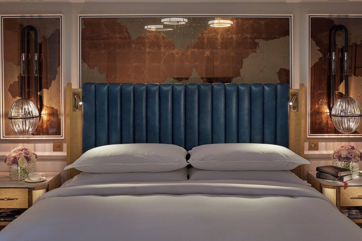 Un grand lit avec une tête de lit rembourrée bleue, deux tables de chevet avec des lampes et des œuvres d’art accrochées aux murs.