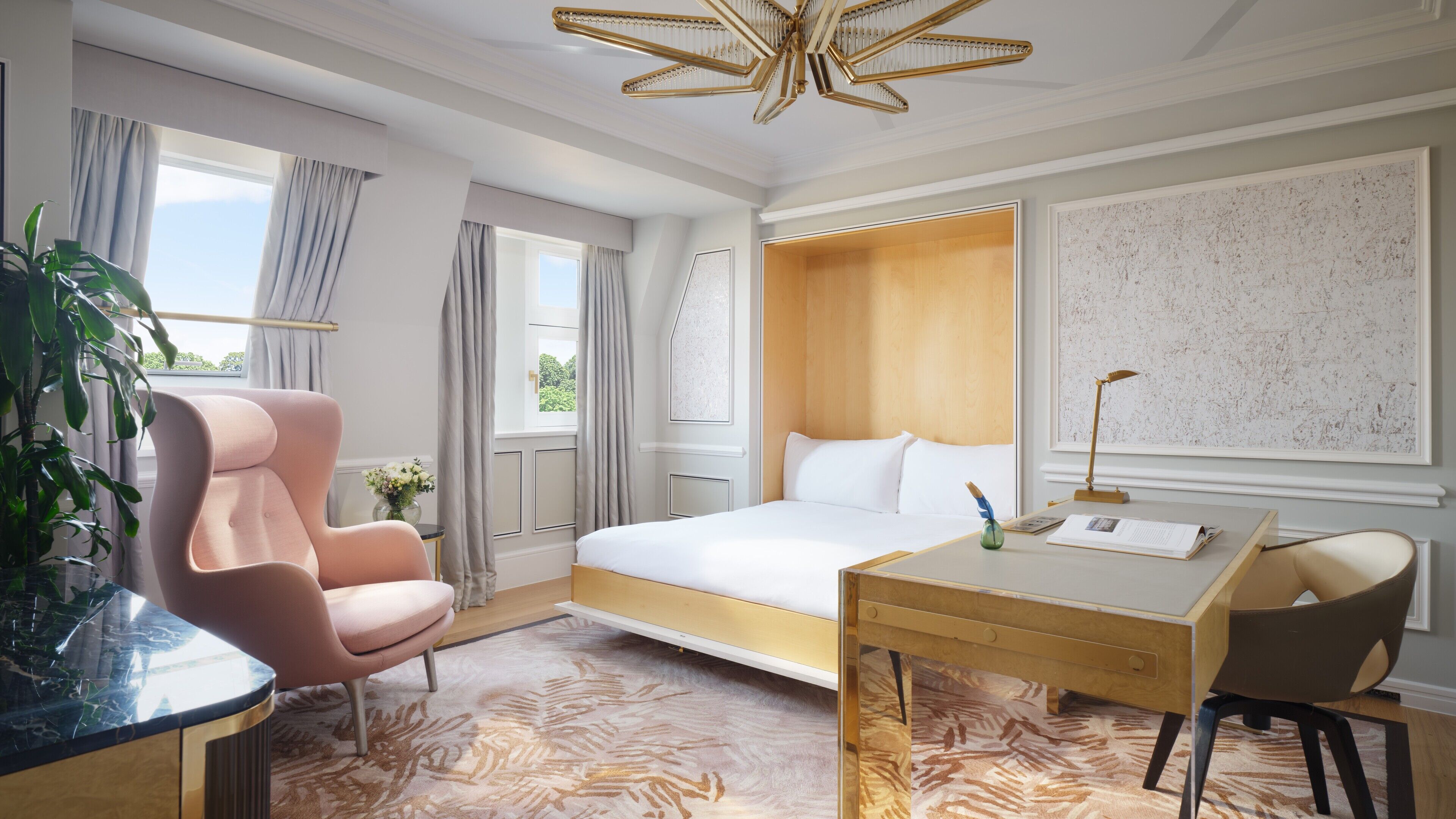 A hotel room with a bed, a desk, a chair, a desk lamp, and a plant.