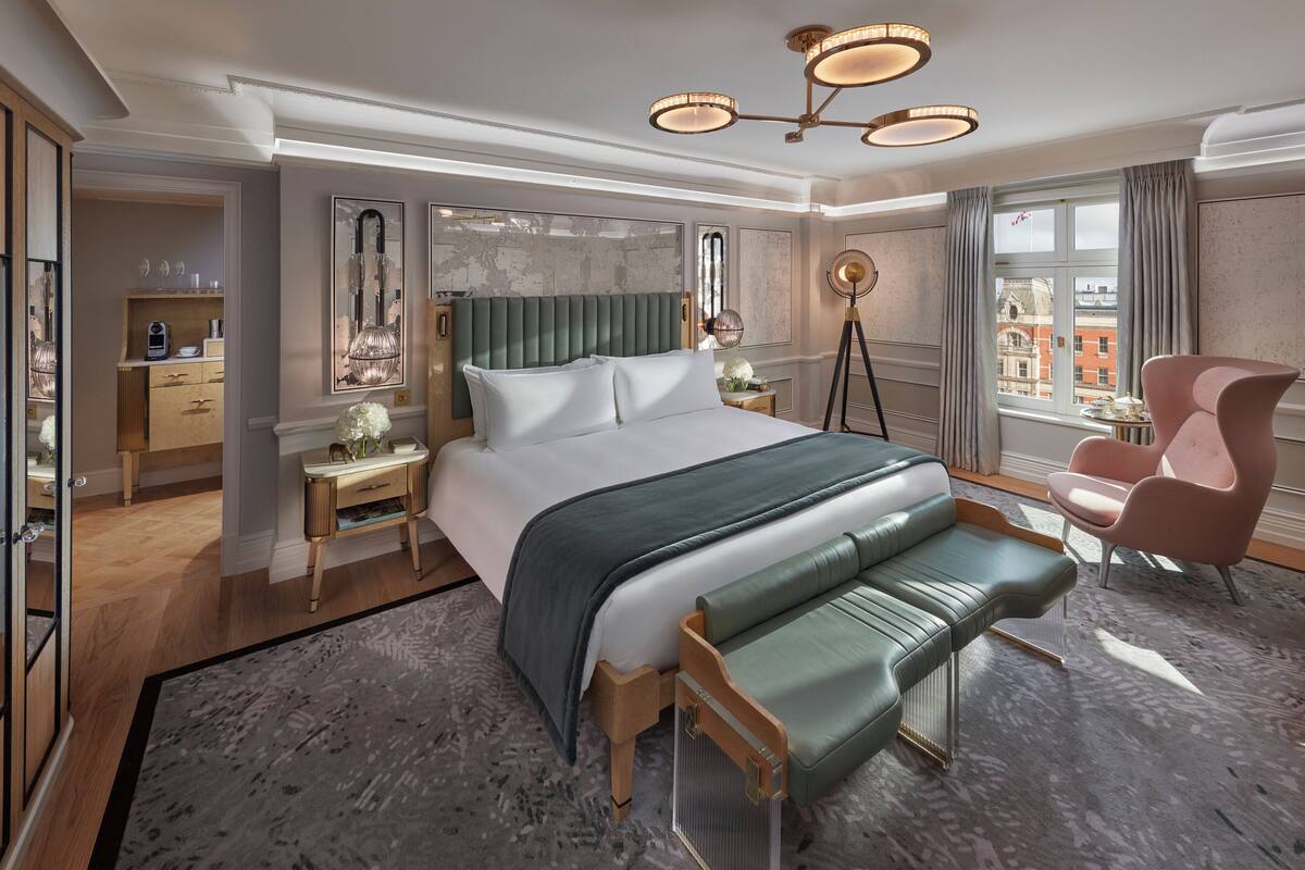 Una habitación de hotel moderna con una cama grande, un banco tapizado de color verde, un sillón rosa y vistas de la ciudad a través de la ventana.
