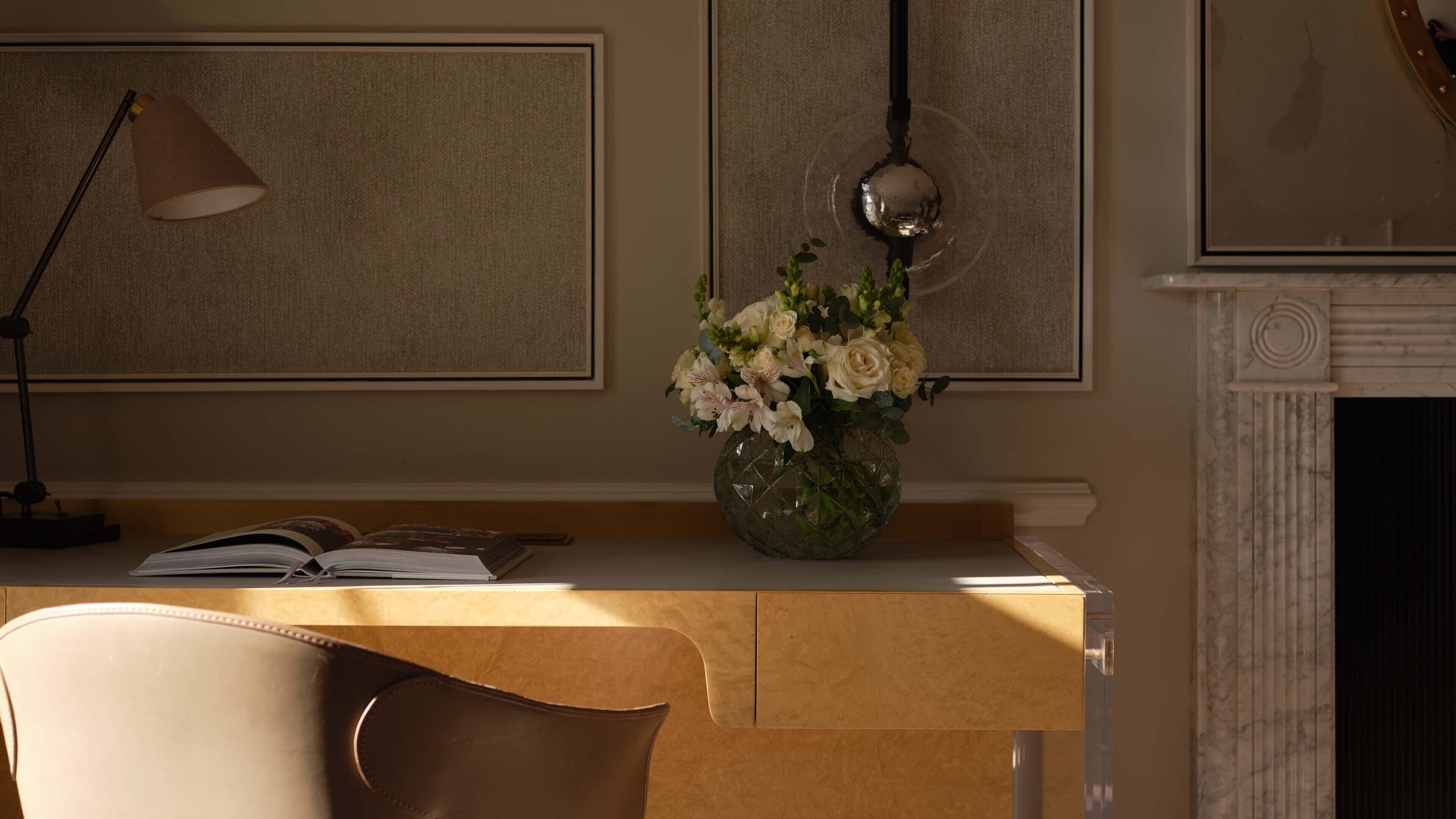A well-lit room with a desk, a lamp, a vase with flowers, and a fireplace.