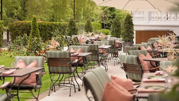 An outdoor dining area with tables and chairs, surrounded by greenery and a hedge wall.