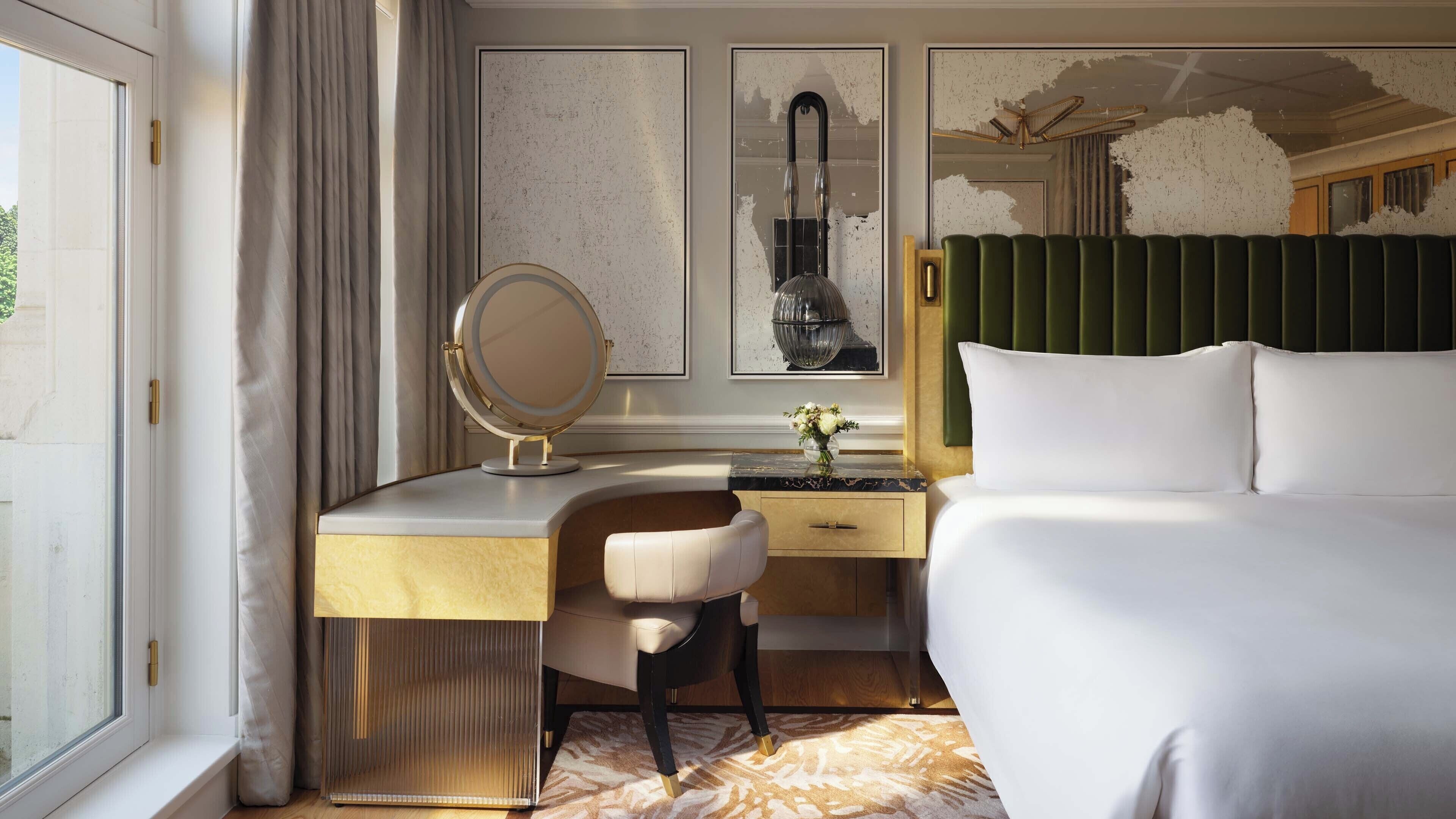 A hotel room with a bed, a desk with a chair, a vanity with a mirror, and a green headboard.