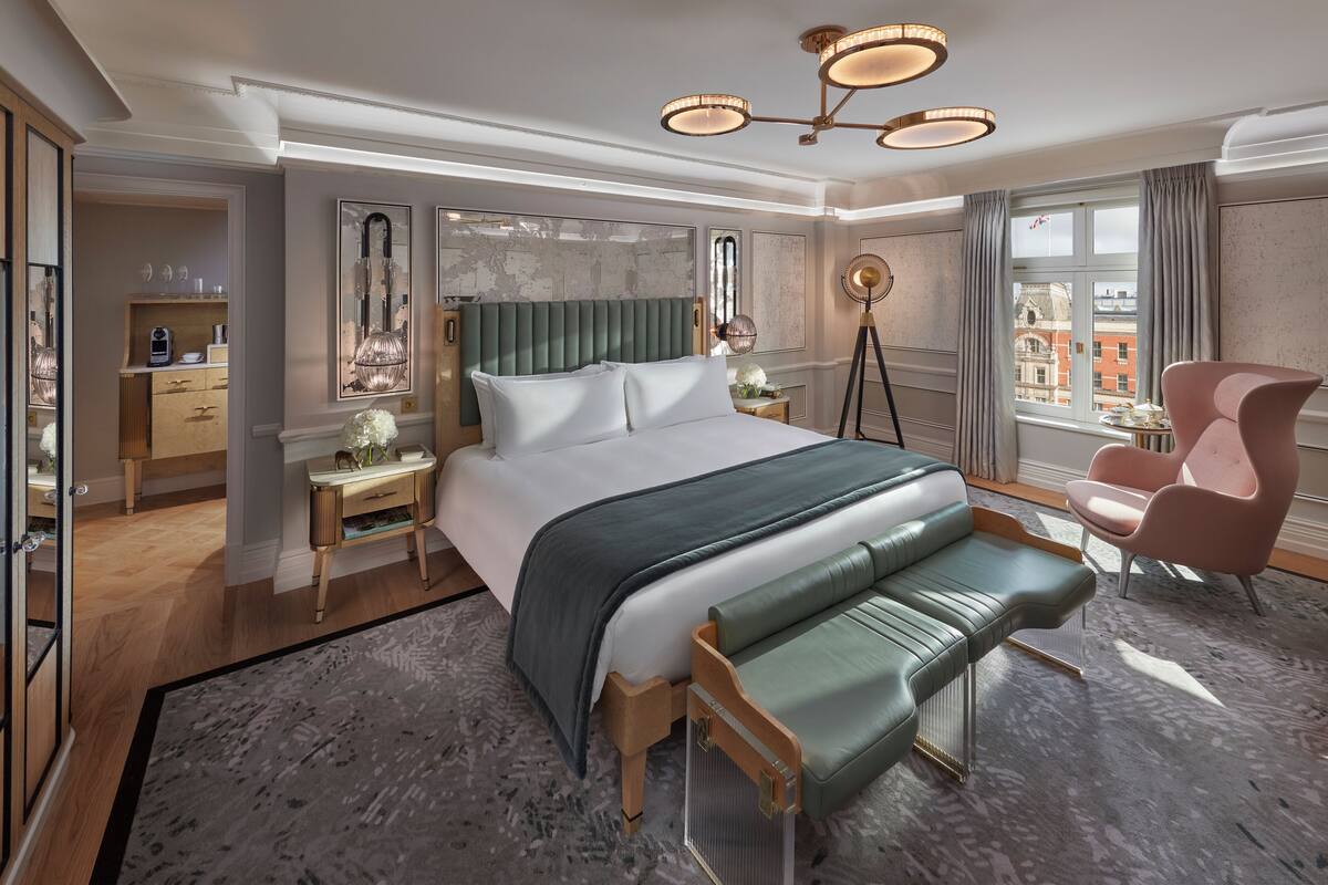 Ein Hotelzimmer mit einem großen Bett, einem grünen Sofa, einem pinken Sessel, einem Nachttisch, einer Lampe und Blick auf die Stadt.