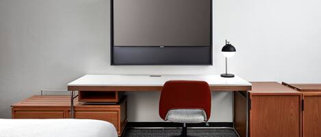 A hotel room with a bed, a desk with a chair, a TV, and a lamp.
